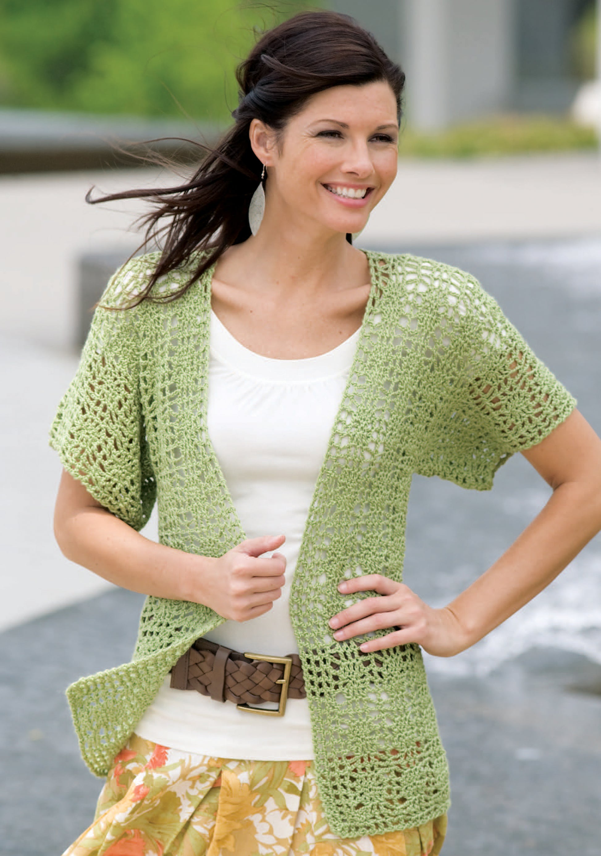 A woman models a light green crochet lace cardigan made with Caron Spa yarn, featured in Leisure Arts’ “Relaxed Fashion—4 Crochet Designs Featuring Caron Spa Yarn.” She pairs it with a white top, brown belt, and floral skirt outdoors.