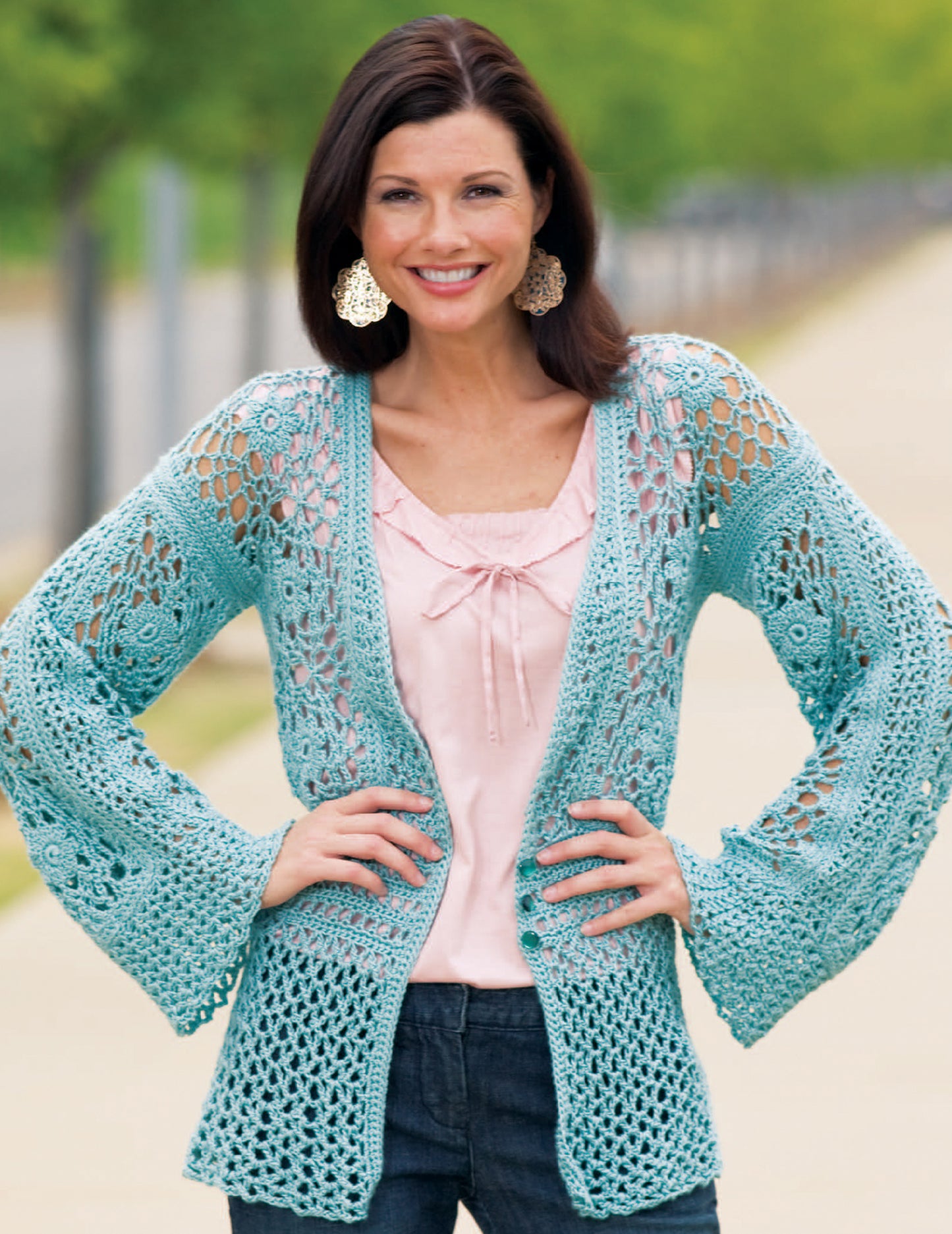 A woman stands outdoors, smiling with her hands on her hips. She wears a feminine crochet lace cardigan made from Caron Spa yarn, created using Relaxed Fashion—4 Crochet Designs by Leisure Arts.