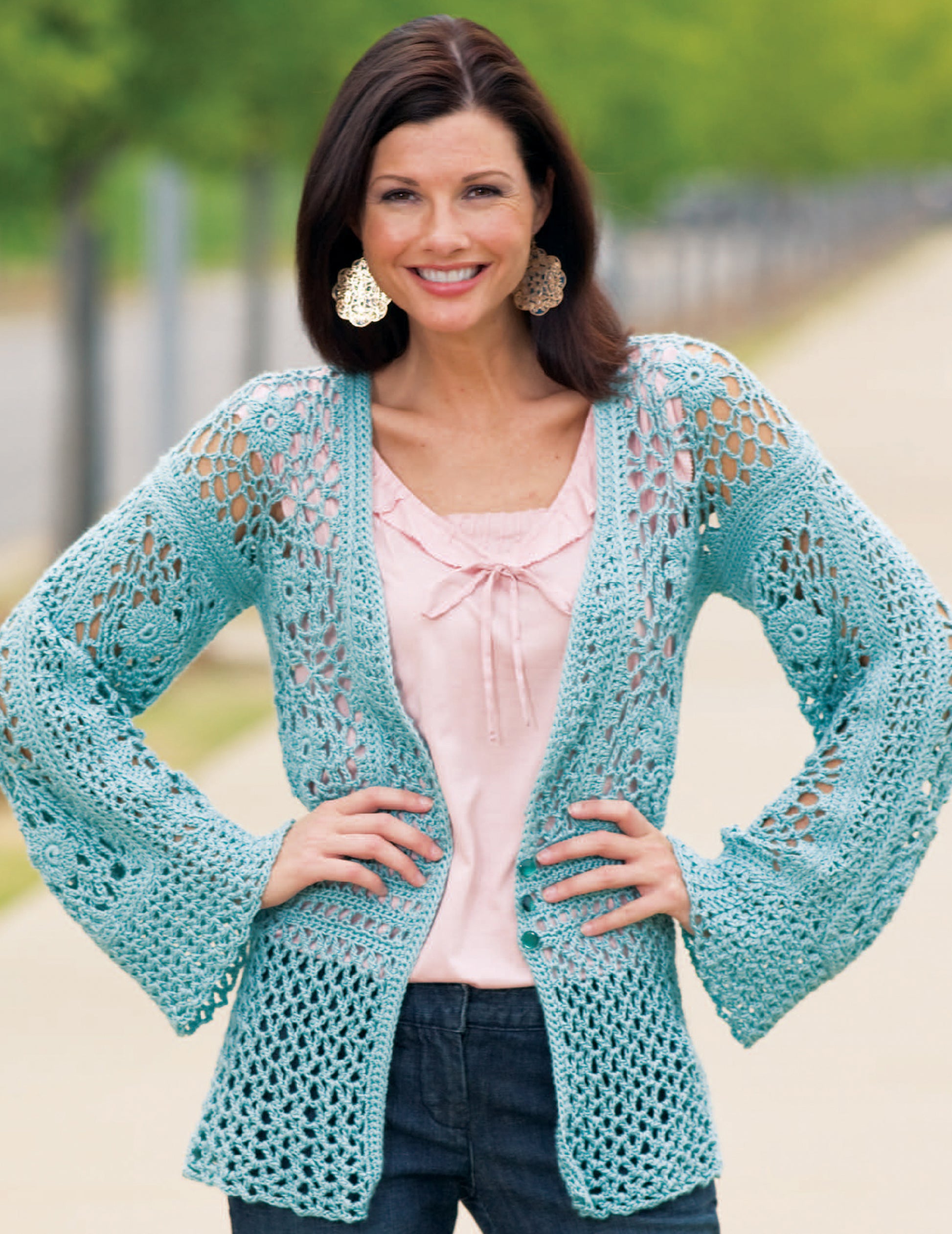 A woman stands outdoors, smiling with her hands on her hips. She wears a feminine crochet lace cardigan made from Caron Spa yarn, created using Relaxed Fashion—4 Crochet Designs by Leisure Arts.