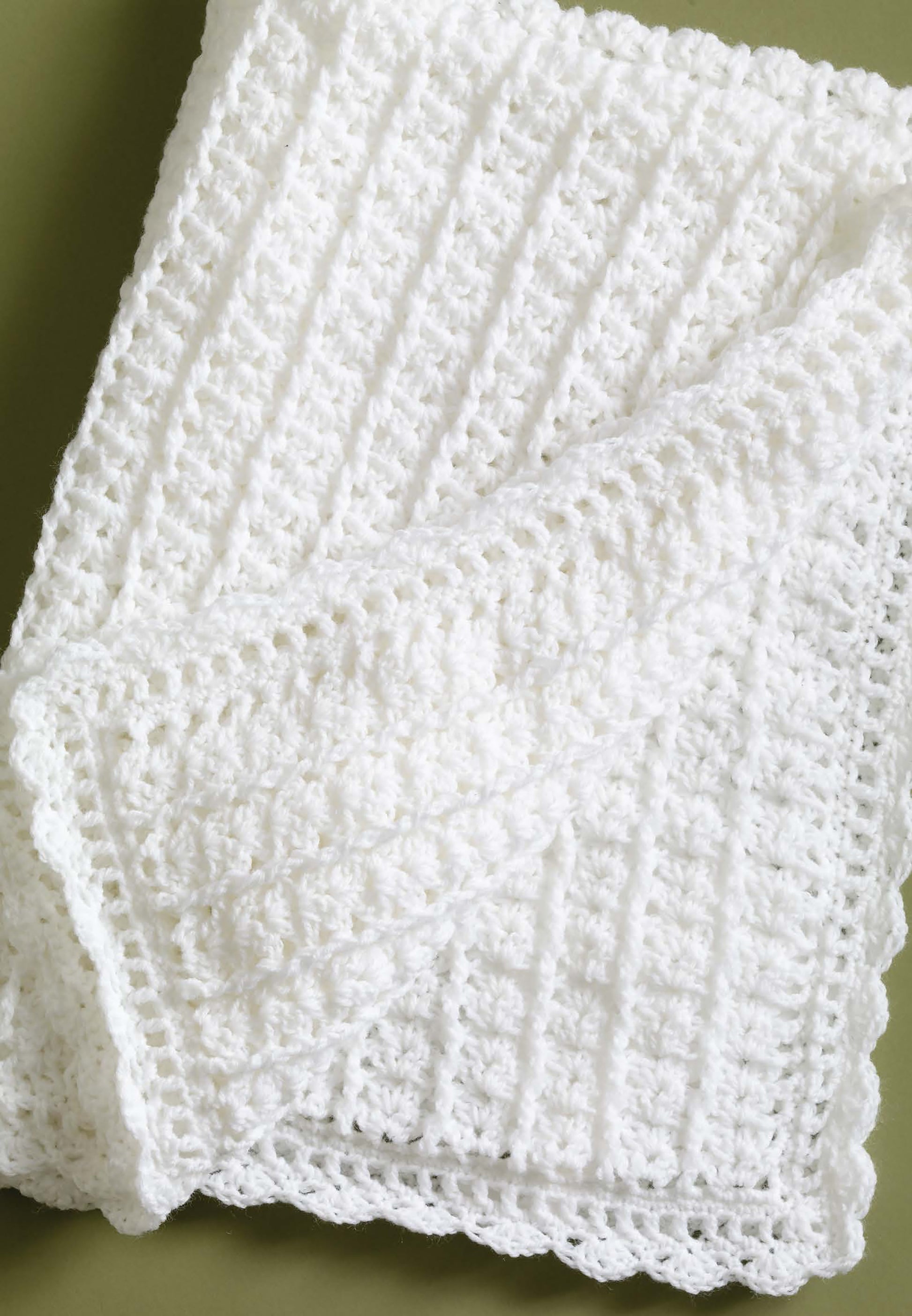 A white crocheted blanket made with lightweight yarn features a ribbed pattern and scalloped edge, shown on green. Pattern from "Small Comforts—4 Crochet & 2 Knit Designs Created with Lion Brand Babysoft" by Leisure Arts.