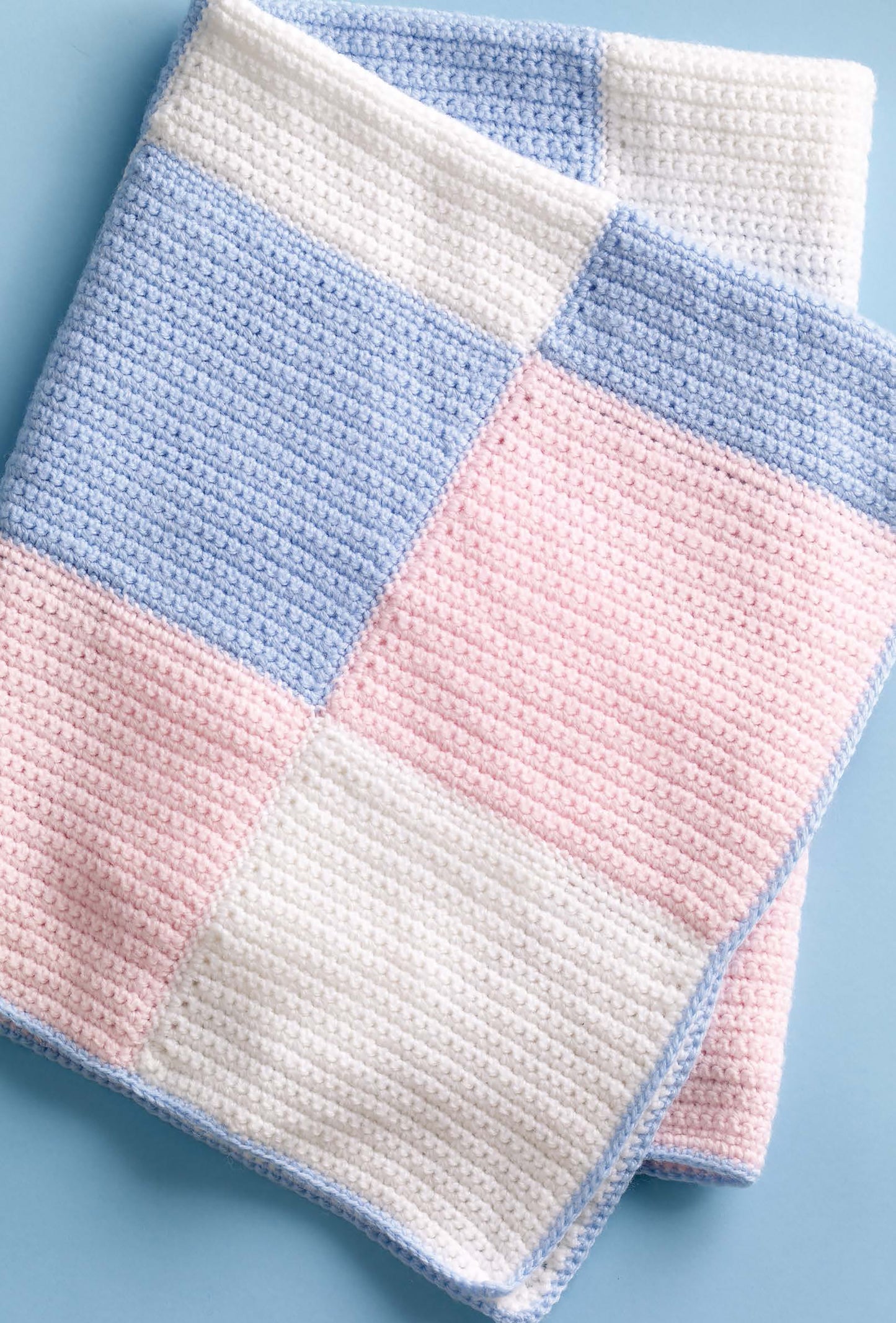 A crocheted blanket made with Lion Brand Babysoft yarn features pastel blue, pink, and white squares. Shown flat on a light blue surface, this cozy look is one of the projects from “Small Comforts” by Leisure Arts.