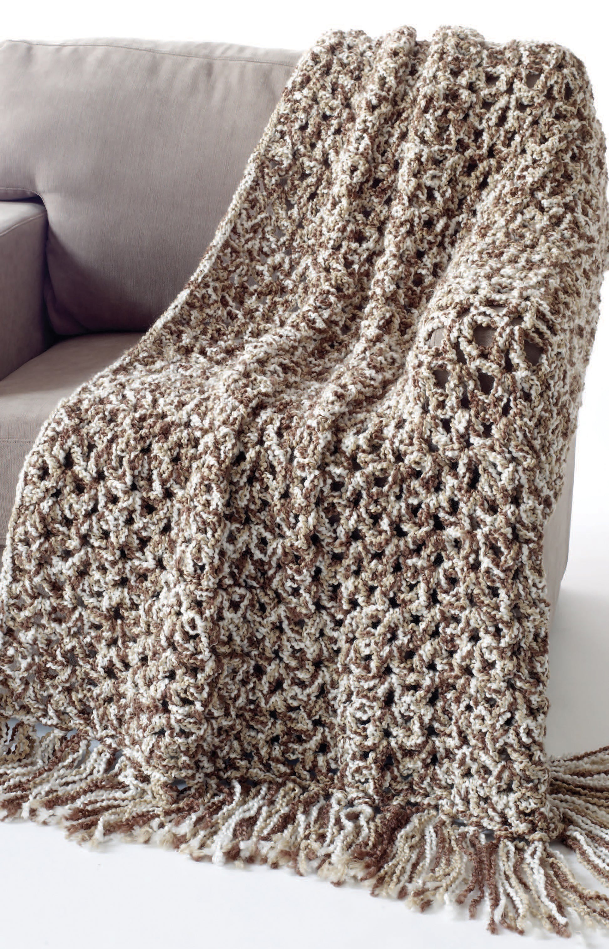 The "Great Warm-Ups" crochet throw by Leisure Arts, featuring a chunky multicolored lacy pattern with fringe, made from bulky yarn, is draped over the arm of a light brown sofa.