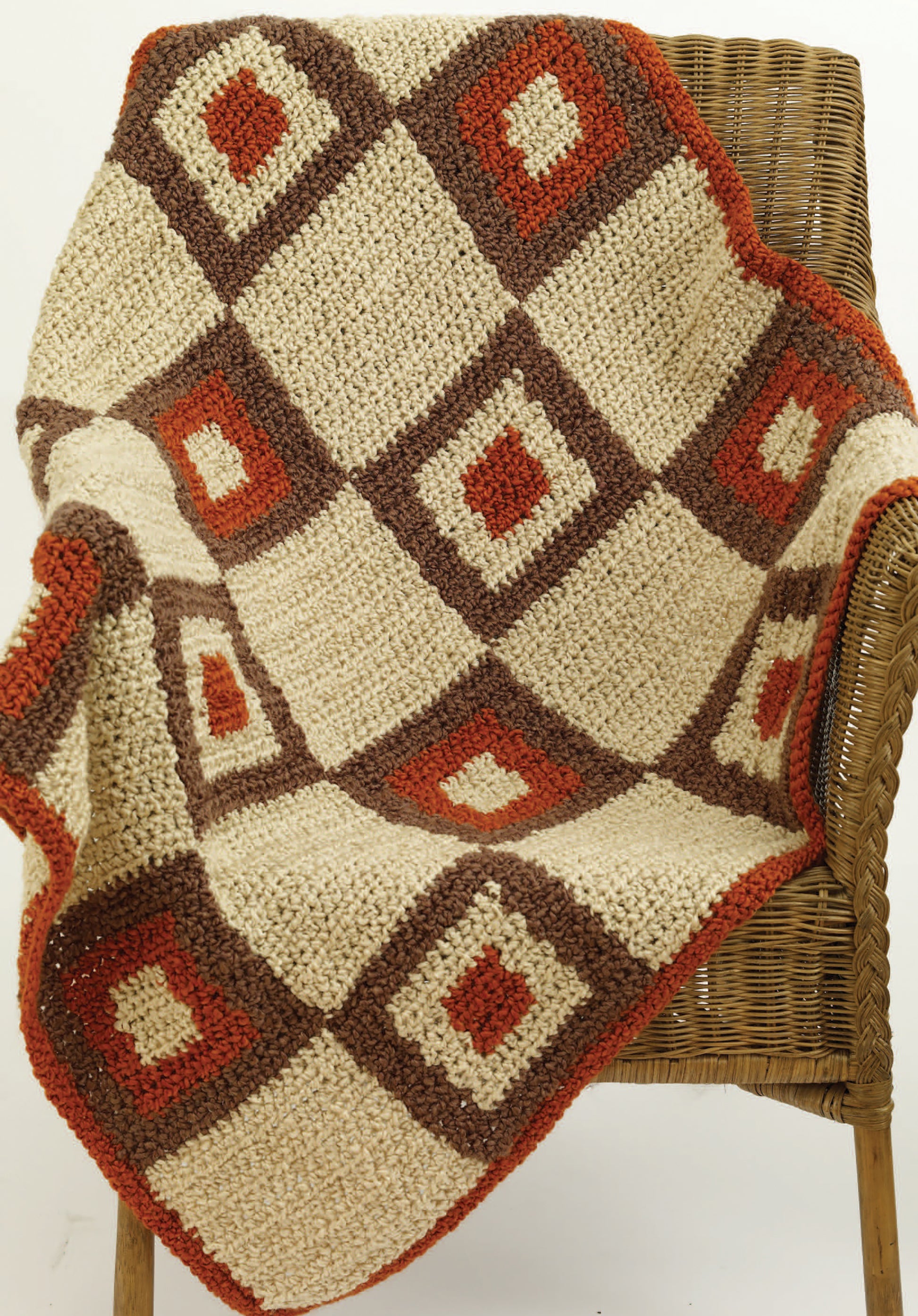 The Great Warm-Ups crochet throw blanket by Leisure Arts features a geometric patchwork of red, brown, and cream diamond squares draped over a wicker chair, inspired by classic afghan designs for cozy comfort.
