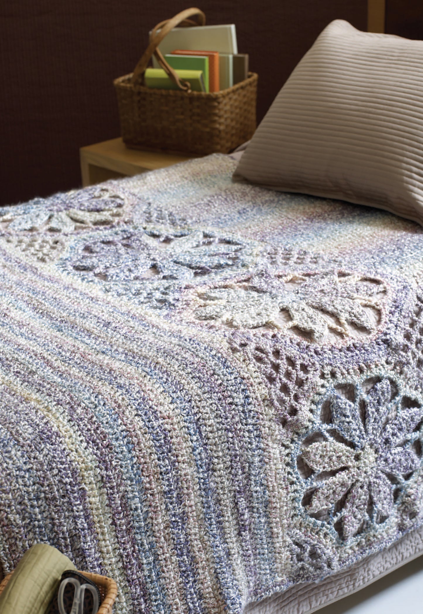 A neatly made bed features the "Great Warm-Ups" crochet throw by Leisure Arts, showcasing pastel stripes and floral patterns in bulky yarn. A beige pillow sits at the head, next to a woven basket of books on a wooden nightstand.