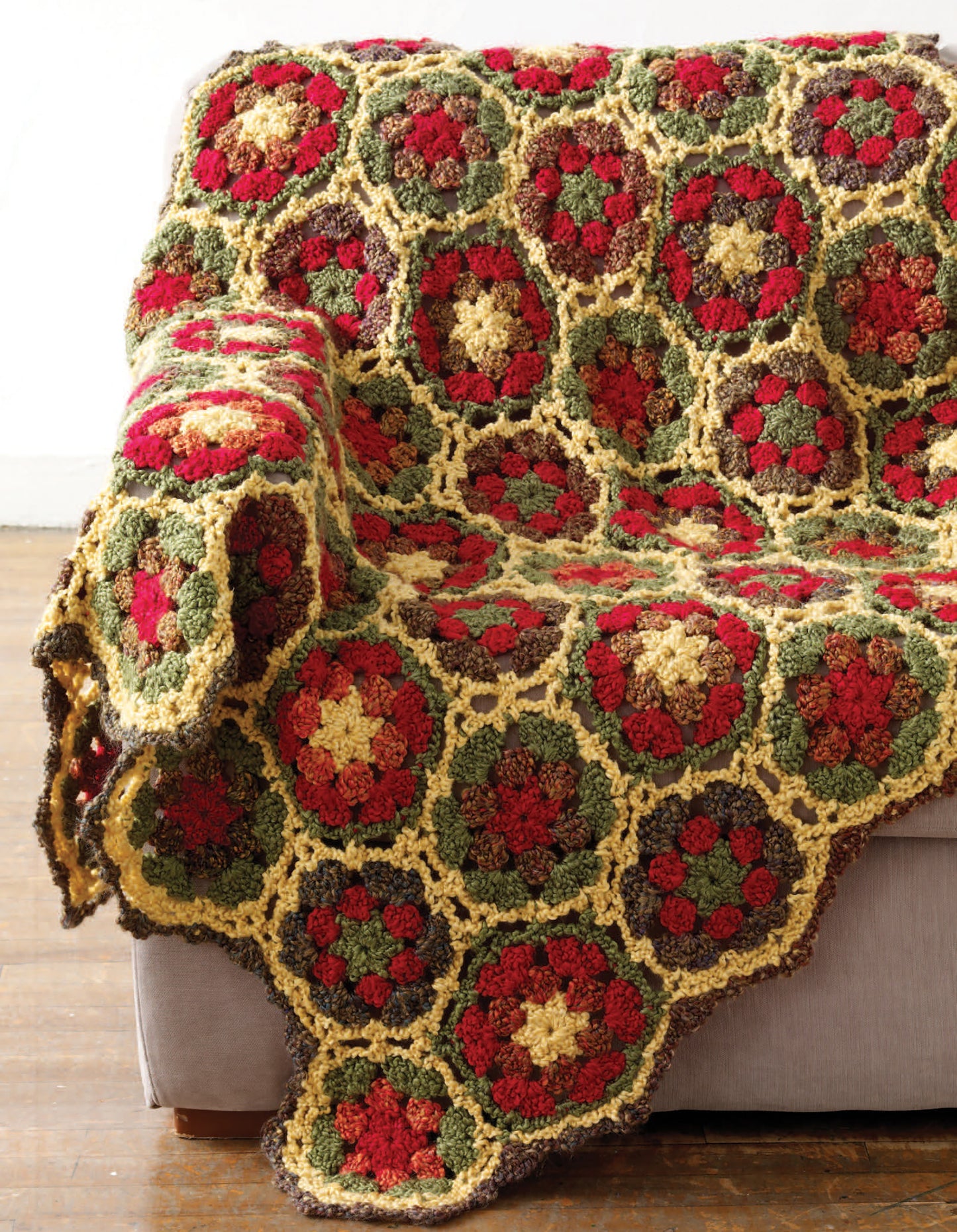 The Great Warm-Ups throw blanket by Leisure Arts, featuring red, green, yellow, and brown hexagonal floral patterns, is draped over the arm and seat of a light gray couch on a wooden floor.