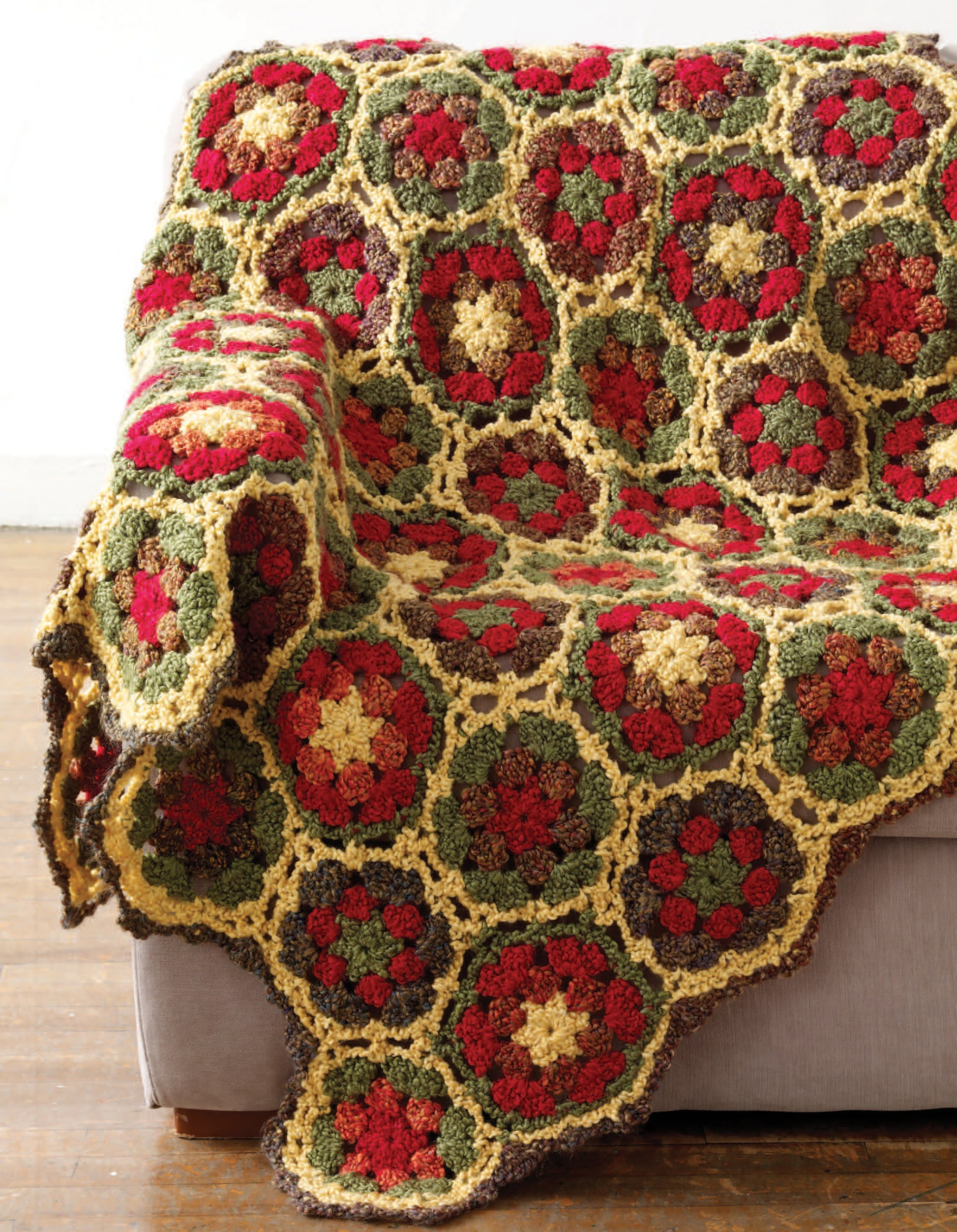 The Great Warm-Ups throw blanket by Leisure Arts, featuring red, green, yellow, and brown hexagonal floral patterns, is draped over the arm and seat of a light gray couch on a wooden floor.