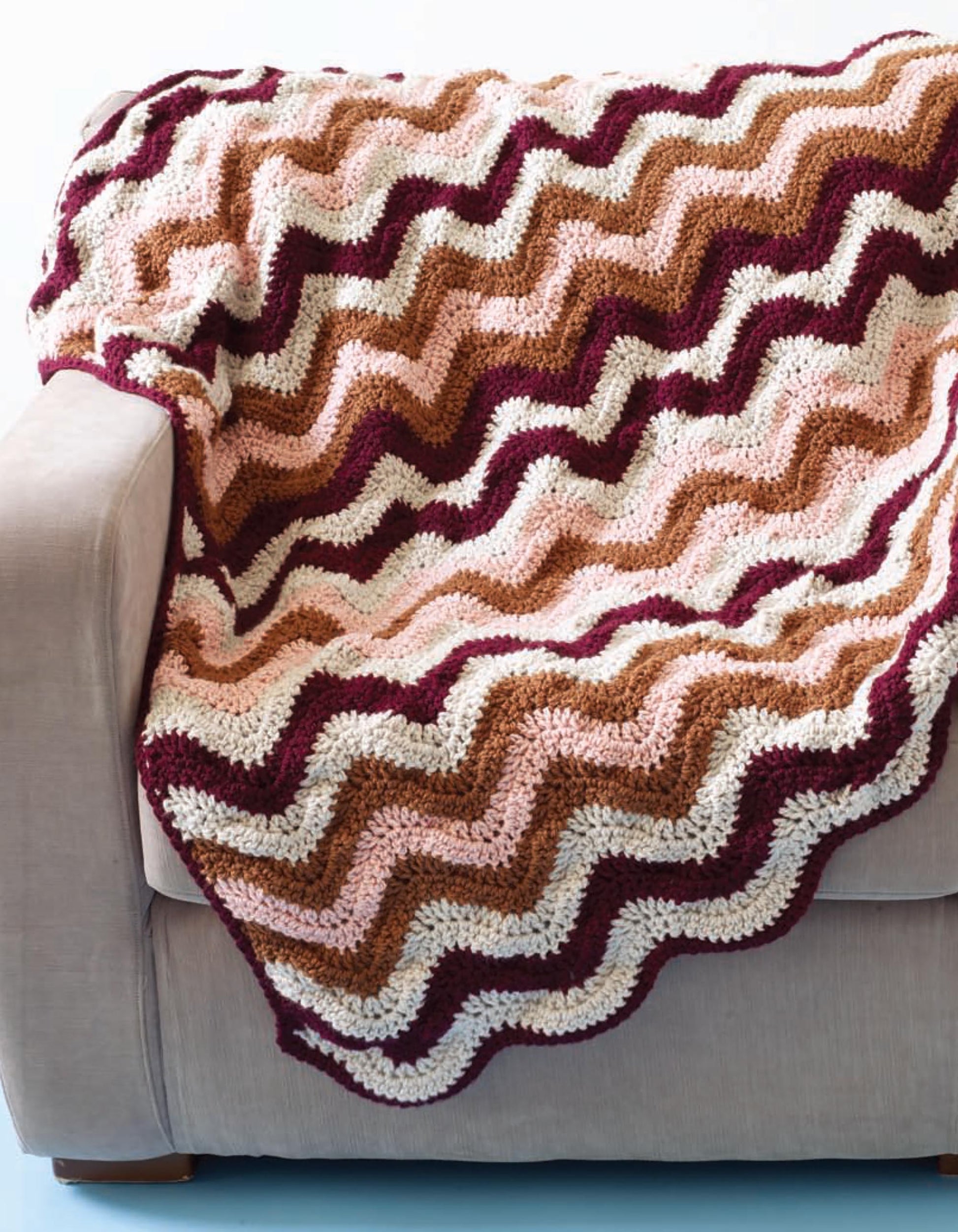 A cozy crocheted blanket made with easy-care yarn from Leisure Arts' Vanna's Fashions of Knit & Crochet, featuring a wavy zigzag pattern in burgundy, cream, brown, and peach, draped on a beige sofa.
