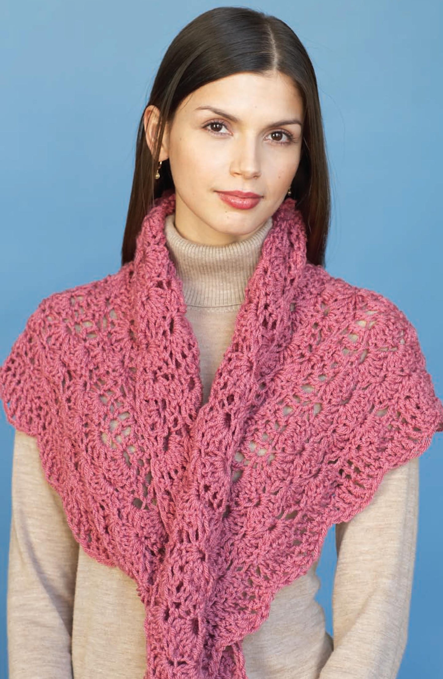 A woman with straight brown hair in a beige turtleneck and chunky pink shawl—knitted from Leisure Arts’ Vanna's Fashions of Knit & Crochet—stands against a solid blue background.