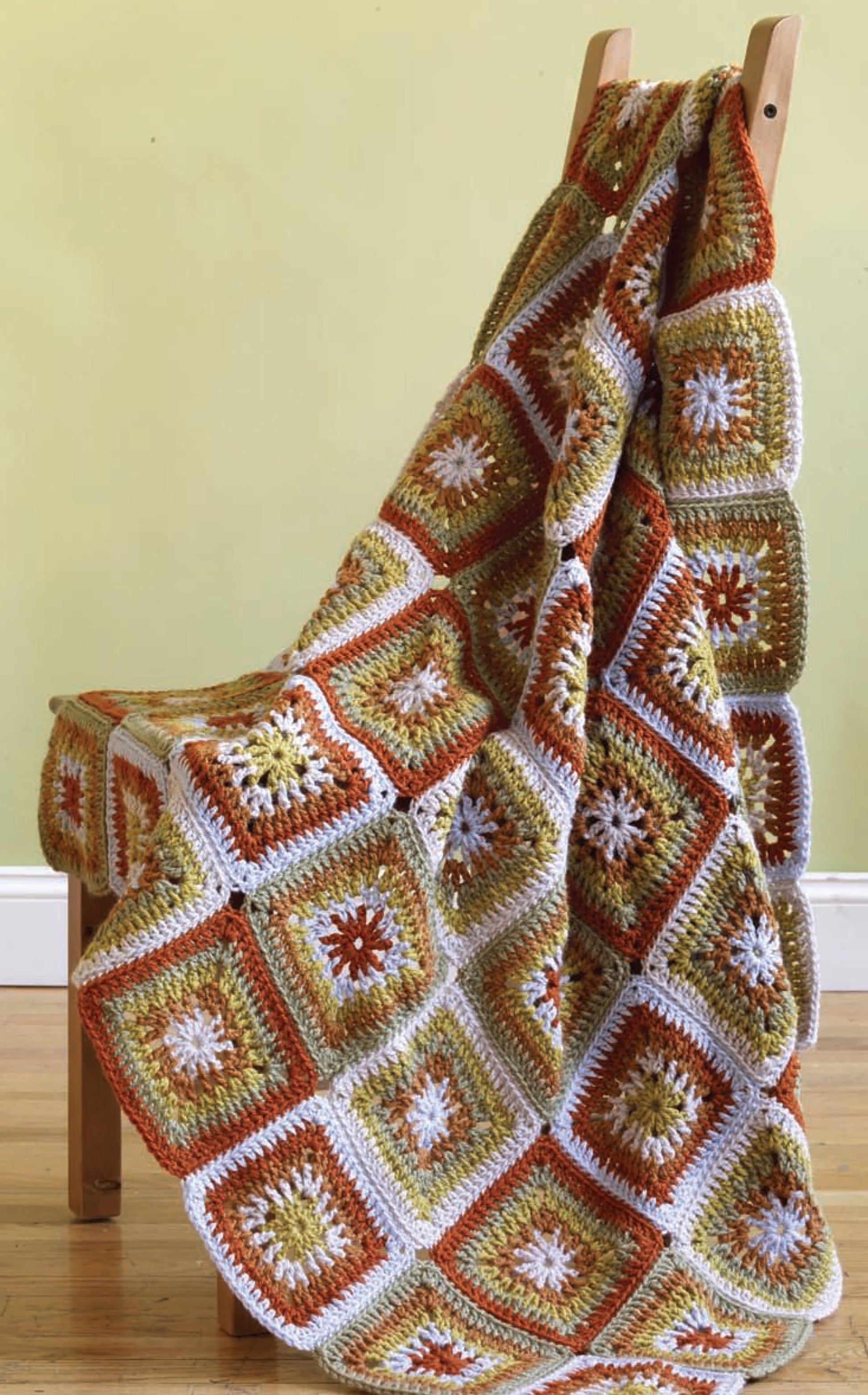 A cozy crocheted blanket made with easy-care yarn from Vanna's Fashions of Knit & Crochet by Leisure Arts, with square patterns in orange, green, white, and yellow, drapes over a wooden chair beside a light yellow wall and wooden floor.