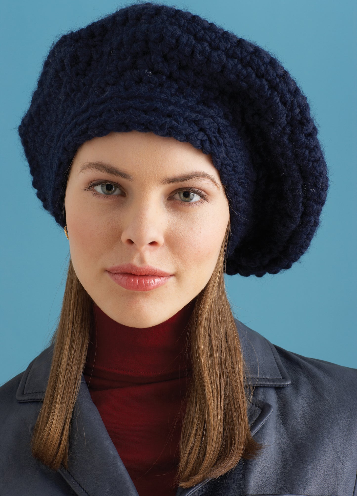 A person with straight brown hair models a navy blue beret made with Lion Brand Wool-Ease Thick & Quick yarn, highlighting designs from Easy & Quick—4 Knit & 2 Crochet Designs by Leisure Arts, against a light blue background.