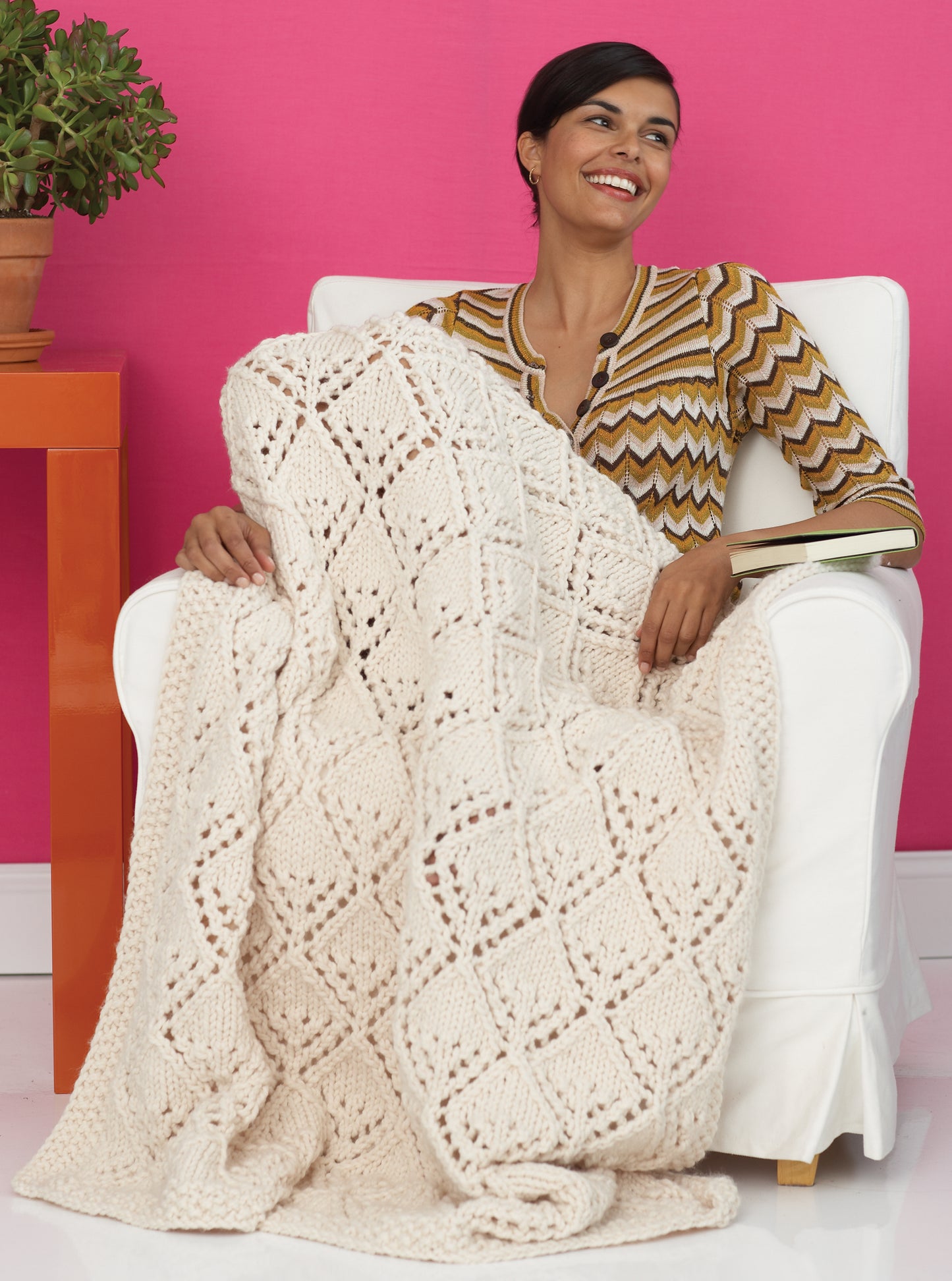A smiling woman sits in a white armchair with a cozy, cream-colored blanket made from Leisure Arts’ Easy & Quick—4 Knit & 2 Crochet Designs (using Lion Brand Wool-Ease Thick & Quick), perfect for beginners. A potted plant sits nearby against a pink wall.