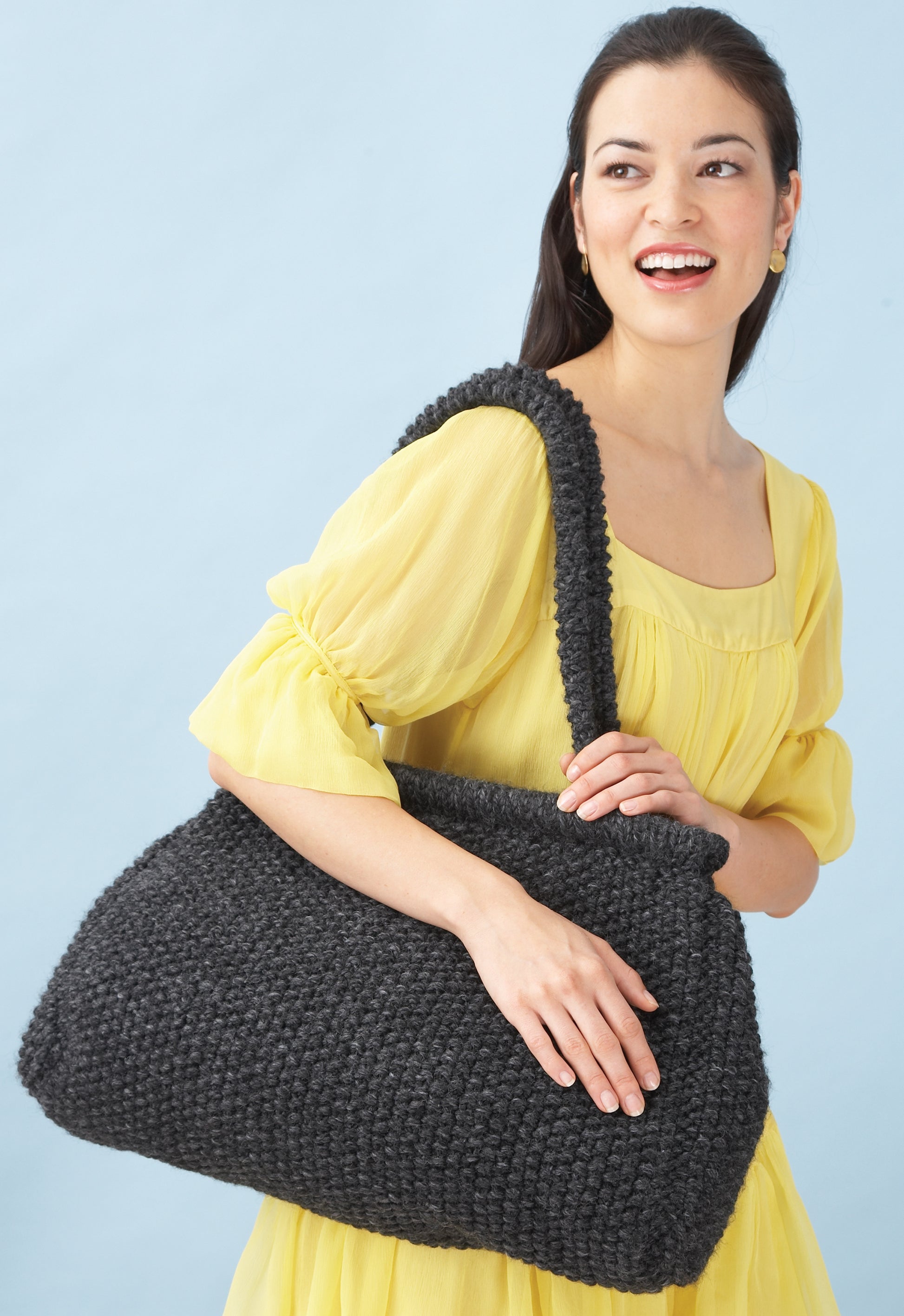 A woman in a yellow dress smiles while holding a large, dark gray bag featuring one of the 4 knit or 2 crochet designs from "Easy & Quick" by Leisure Arts, made with Lion Brand Wool-Ease Thick & Quick yarn, against a light blue background.