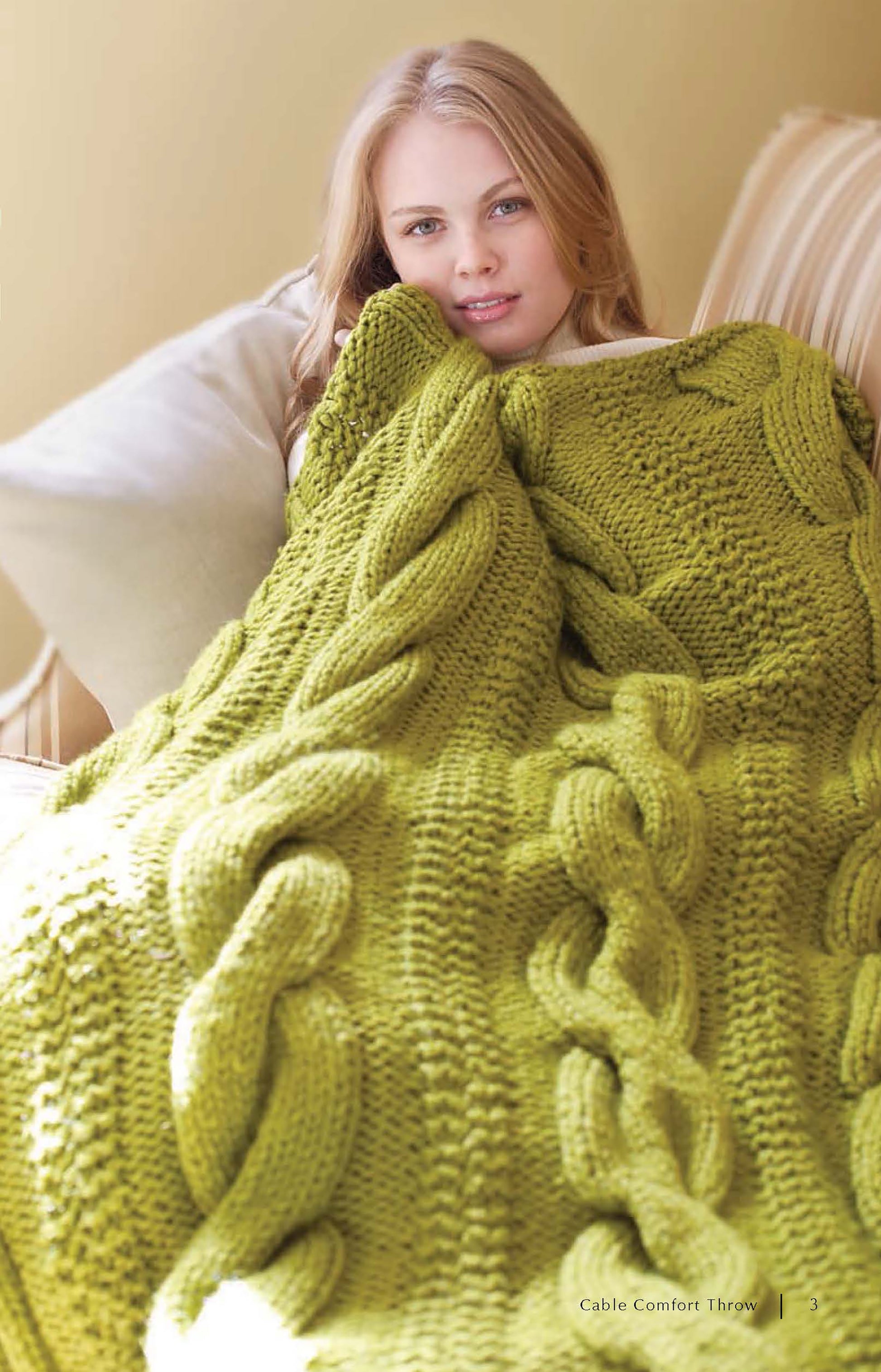 A person with long blonde hair sits on a couch, wrapped in a chunky green cable-knit blanket, looking relaxed and cozy. Get inspired for your next project with "Super Cute Super Quick Crochet" by Leisure Arts.