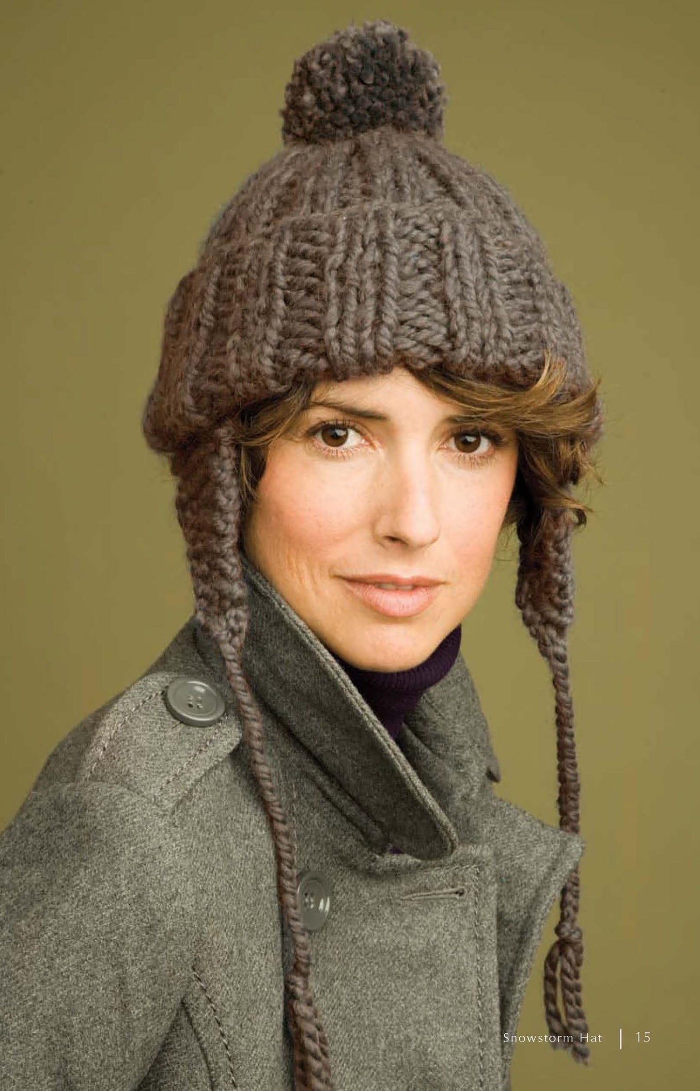 A person in a thick gray knit hat with ear flaps, inspired by patterns from "Super Cute Super Quick Crochet" by Leisure Arts, and a gray coat poses against a plain olive-green background.