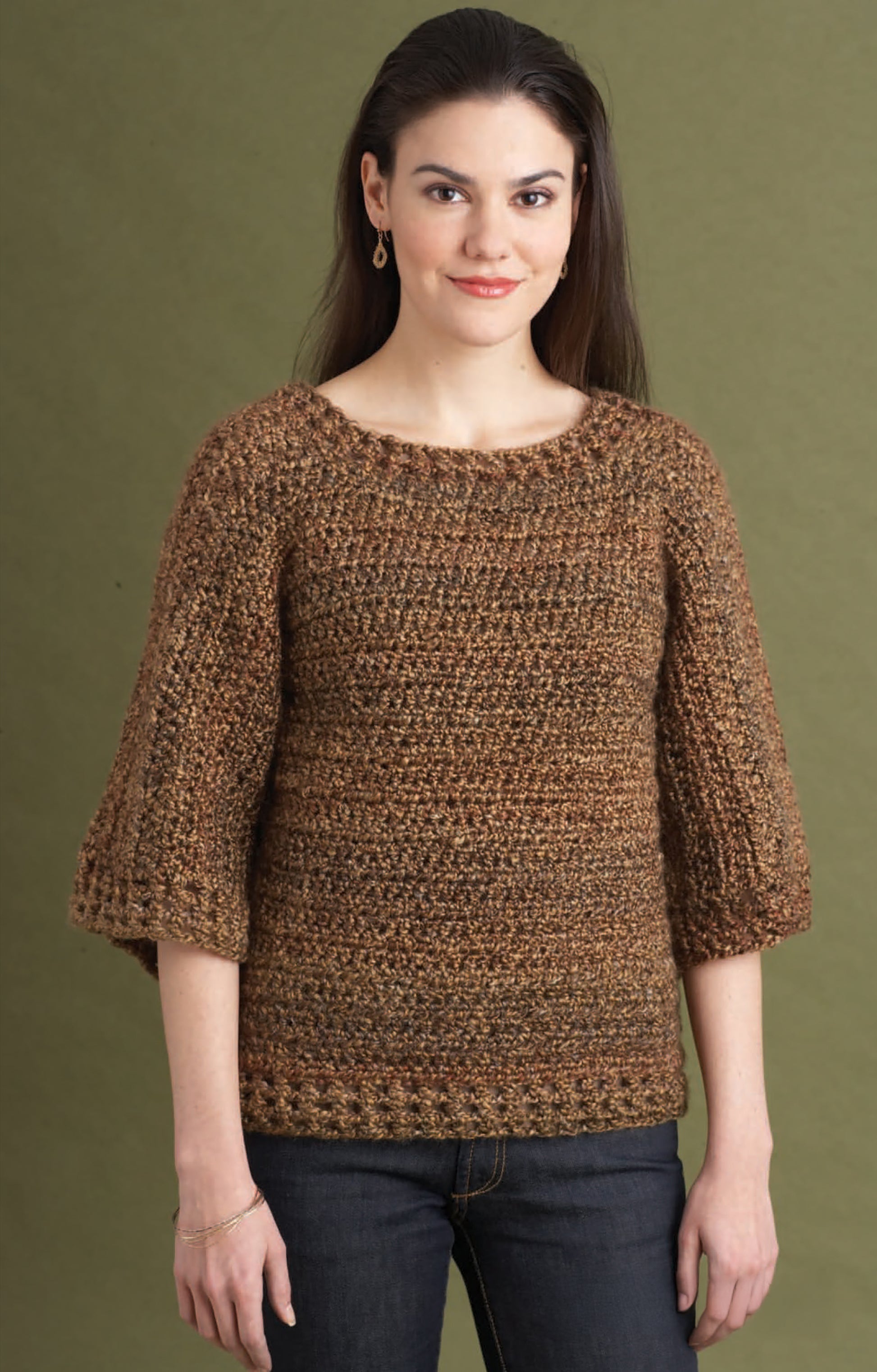 A woman wears the Leisure Arts "Homespun Warmth" brown pullover sweater, crafted with bulky yarn, scoop neckline, and flared three-quarter sleeves, paired with dark jeans against an olive green background.
