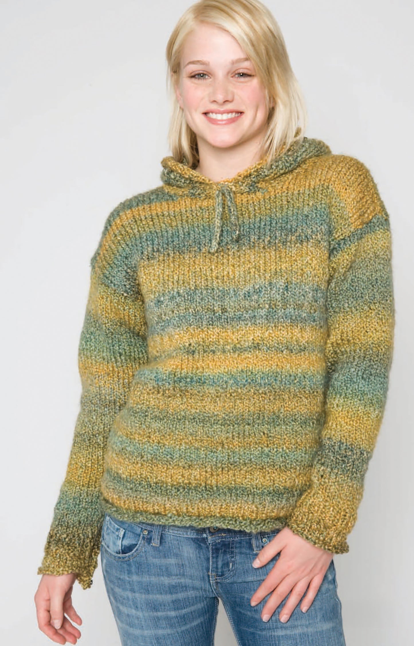 A smiling blonde person wears the Leisure Arts "Homespun Warmth" green and yellow striped bulky knit sweater with blue jeans, standing against a plain light background.