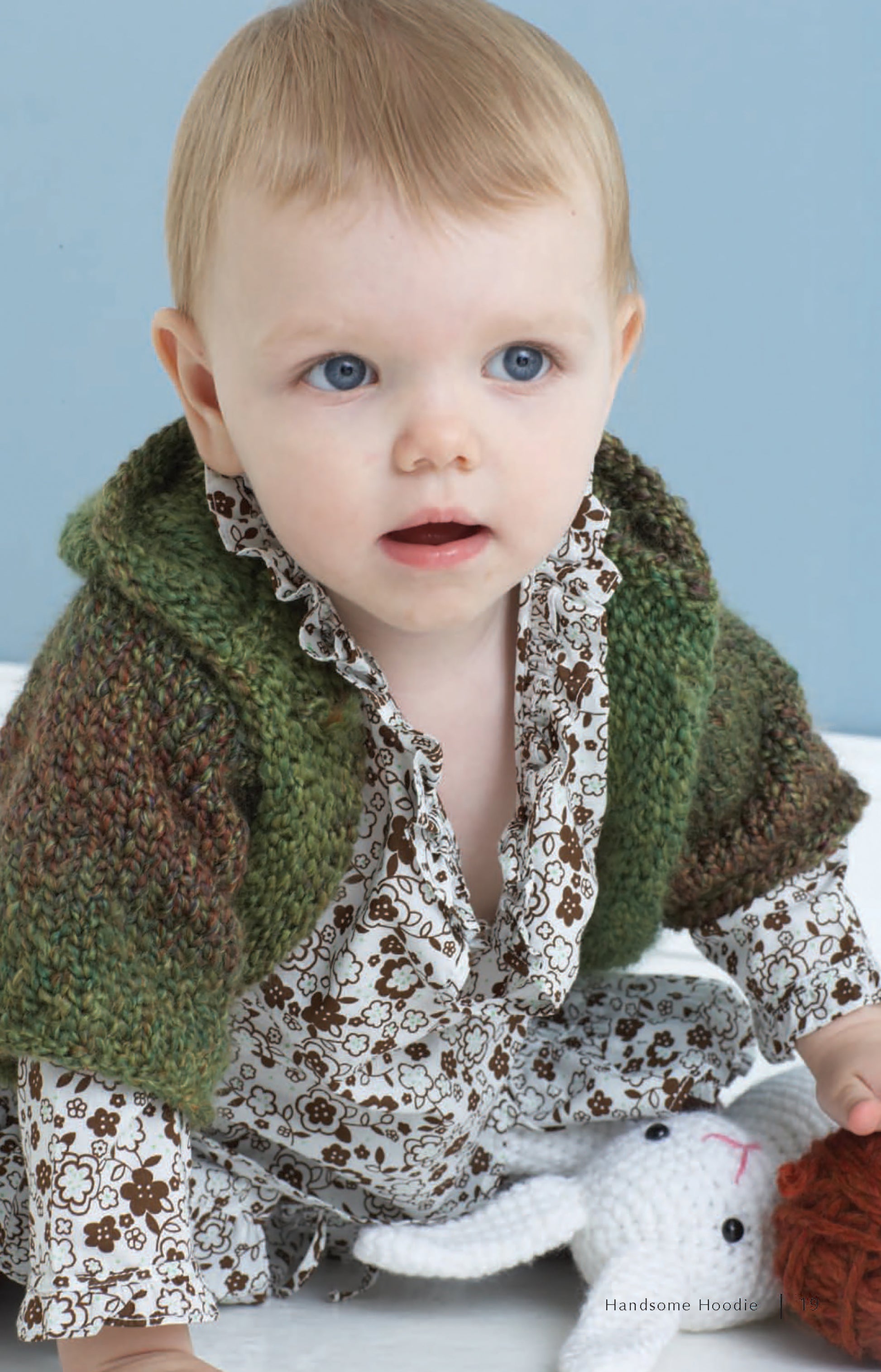 A baby with light brown hair and blue eyes wears a green hoodie made from Leisure Arts’ Homespun Warmth bulky yarn, paired with a floral outfit, while sitting on the floor and holding a white stuffed animal against a light blue background.