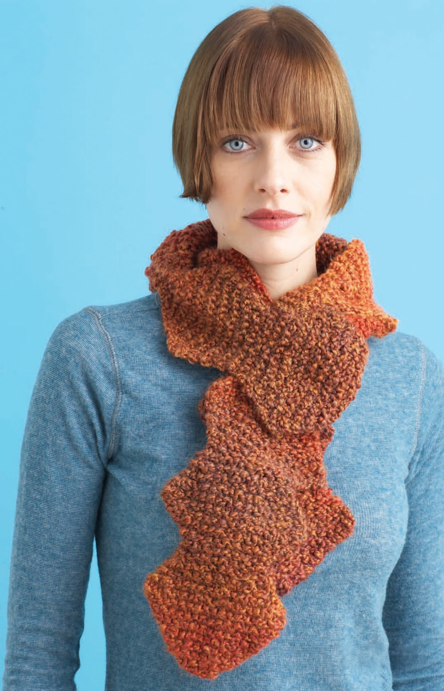 A woman with short brown hair wears a blue sweater and the Homespun Warmth scarf by Leisure Arts, featuring a wavy, layered rust-colored design made from bulky weight yarn, standing in front of a light blue background.