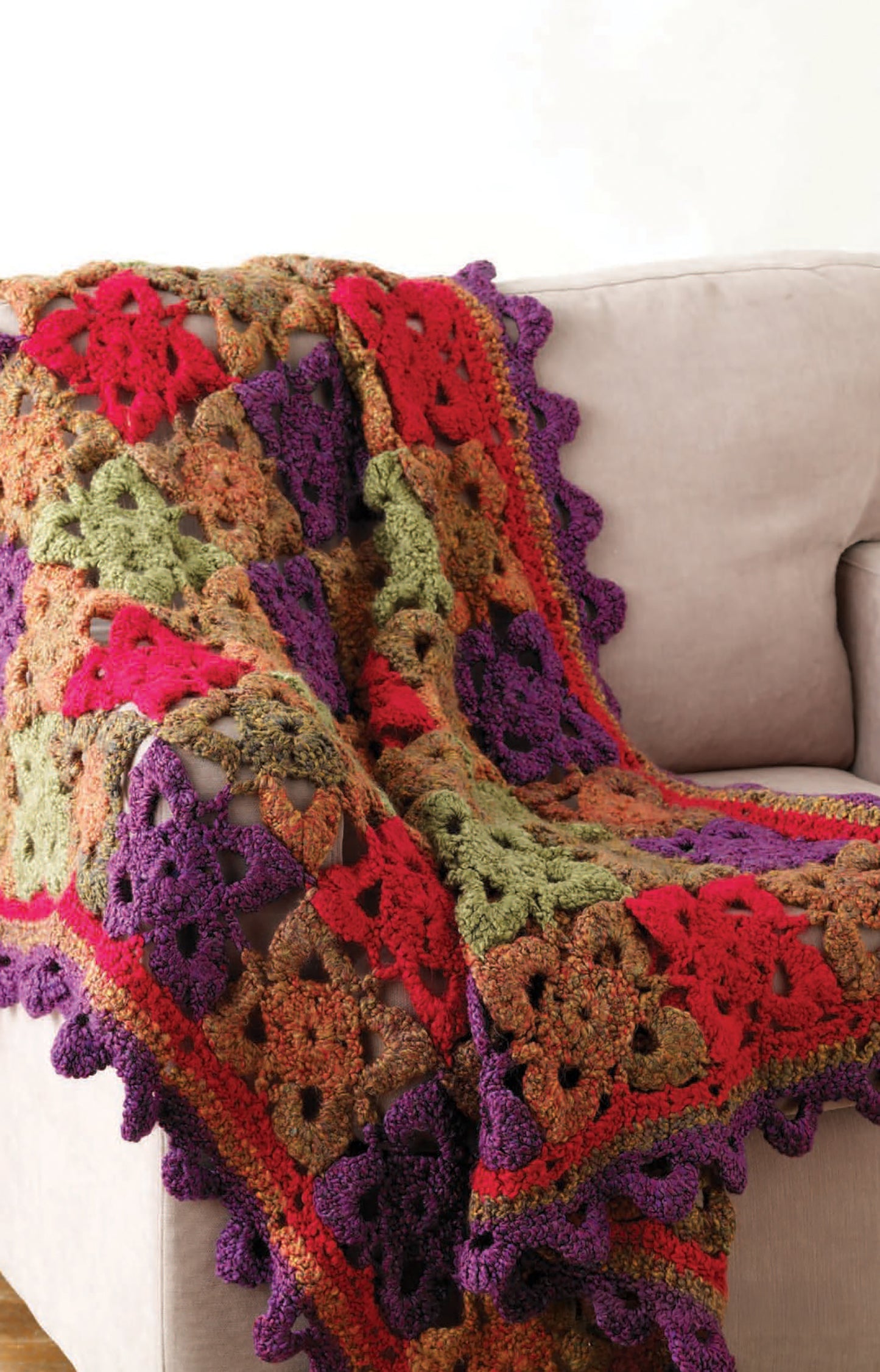 Leisure Arts' Homespun Warmth: a vibrant crochet blanket with bulky yarn red, green, purple, and orange granny squares and a scalloped purple border, draped over a light beige sofa.