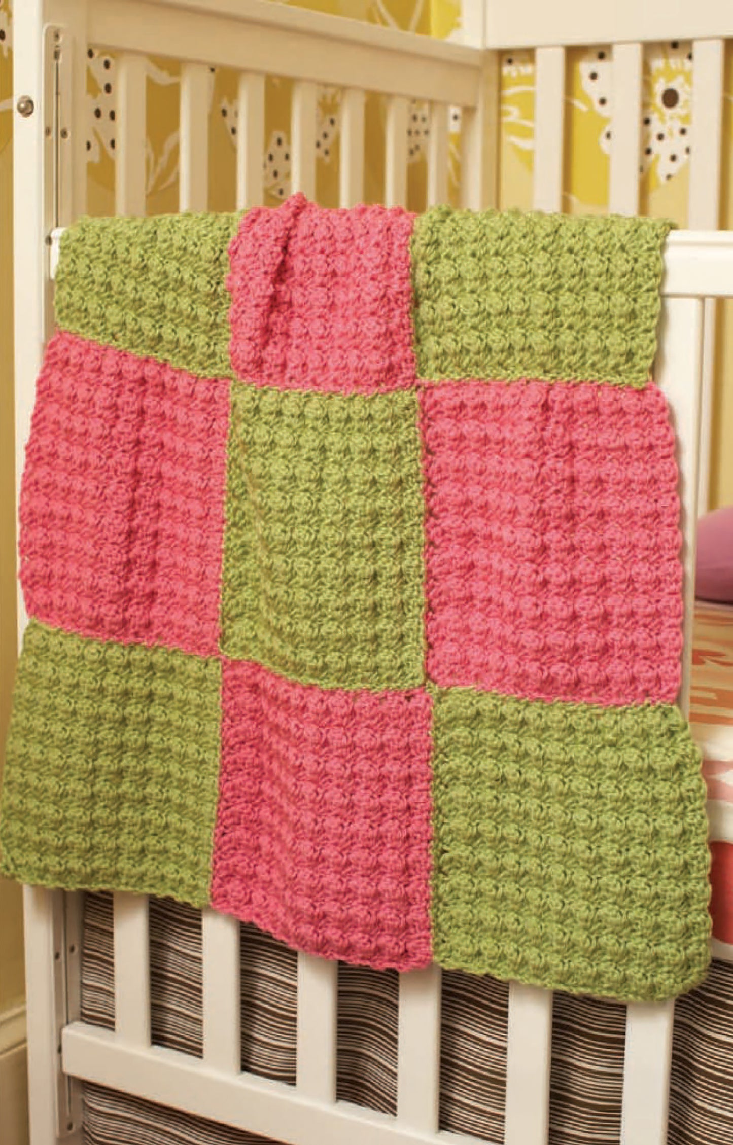 A crocheted blanket with a pink and green checkerboard pattern made from Leisure Arts Vanna's Choice Playful Style yarn hangs over a white baby crib with a striped mattress, set against a yellow patterned wall.