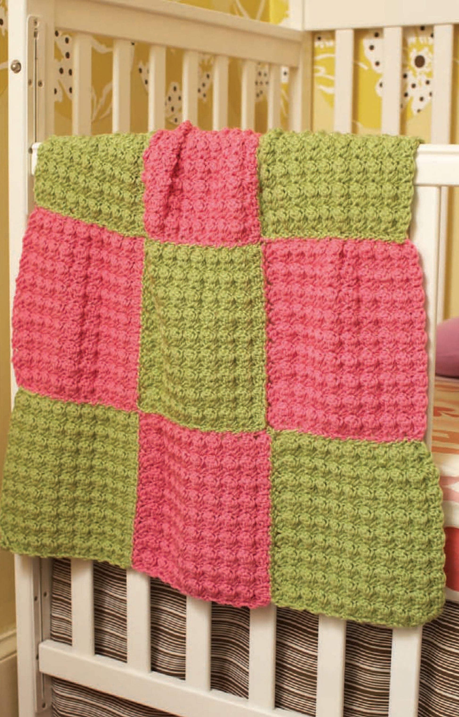 A crocheted blanket with a pink and green checkerboard pattern made from Leisure Arts Vanna's Choice Playful Style yarn hangs over a white baby crib with a striped mattress, set against a yellow patterned wall.