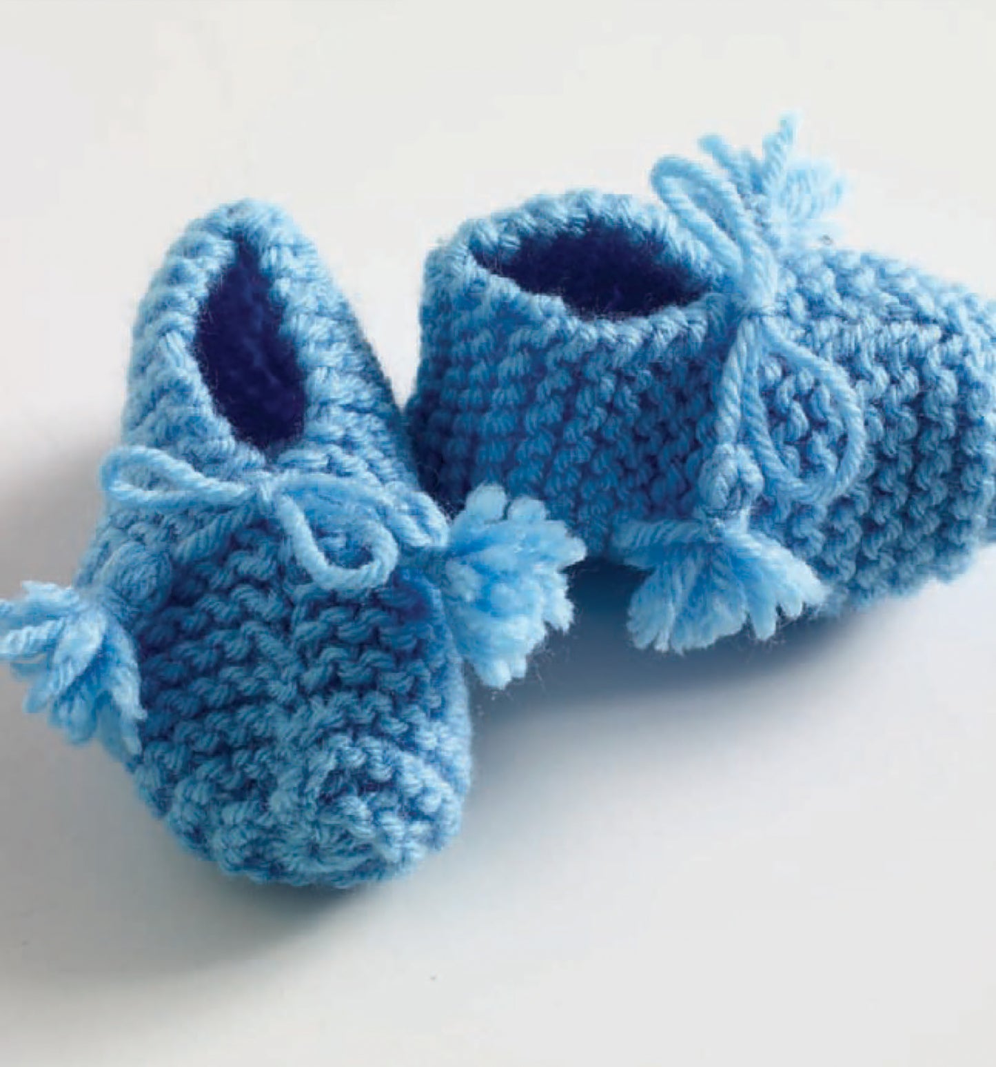 A pair of hand-knitted blue baby booties made with Leisure Arts Vanna's Choice Playful Style yarn, decorated with bows and small tassels, displayed on a white surface.