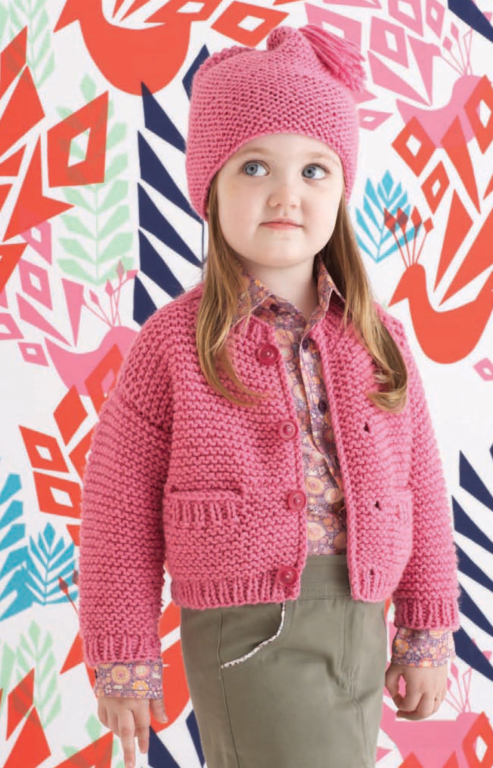 A young girl in a pink knit hat and cardigan made with Leisure Arts’ Vanna's Choice Playful Style yarn stands before a colorful geometric and floral background. She has long brown hair, a patterned shirt, and green pants.