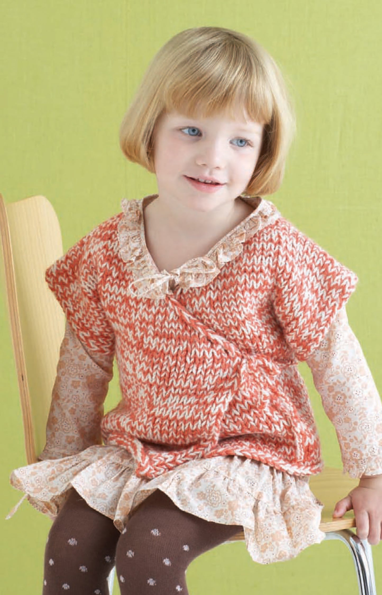 A young child with short blonde hair sits on a wooden chair, wearing a peach and white patterned sweater over a cream dress with brown tights, all made from Leisure Arts Vanna's Choice Playful Style yarn, against a light green background.