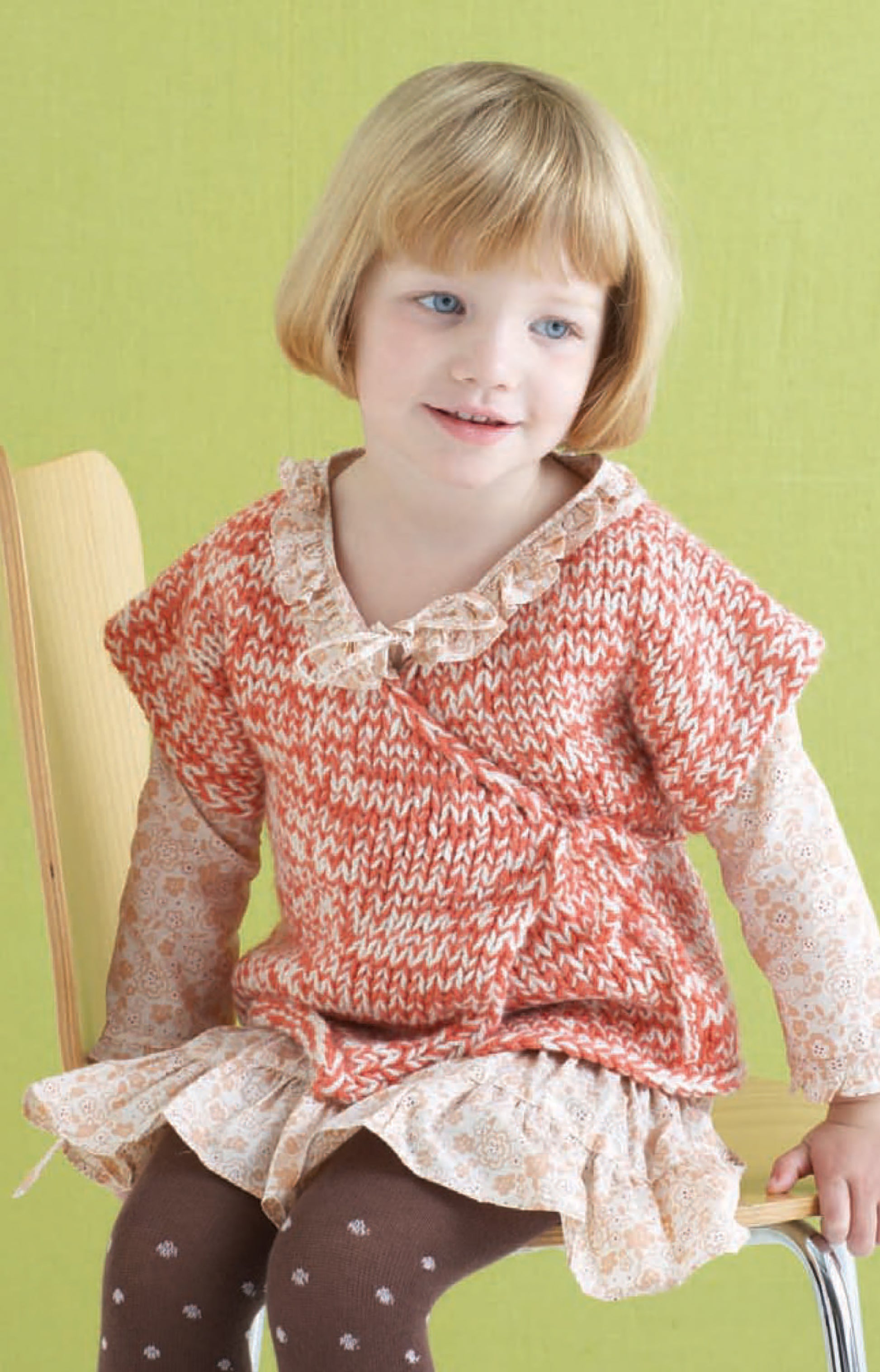 A young child with short blonde hair sits on a wooden chair, wearing a peach and white patterned sweater over a cream dress with brown tights, all made from Leisure Arts Vanna's Choice Playful Style yarn, against a light green background.
