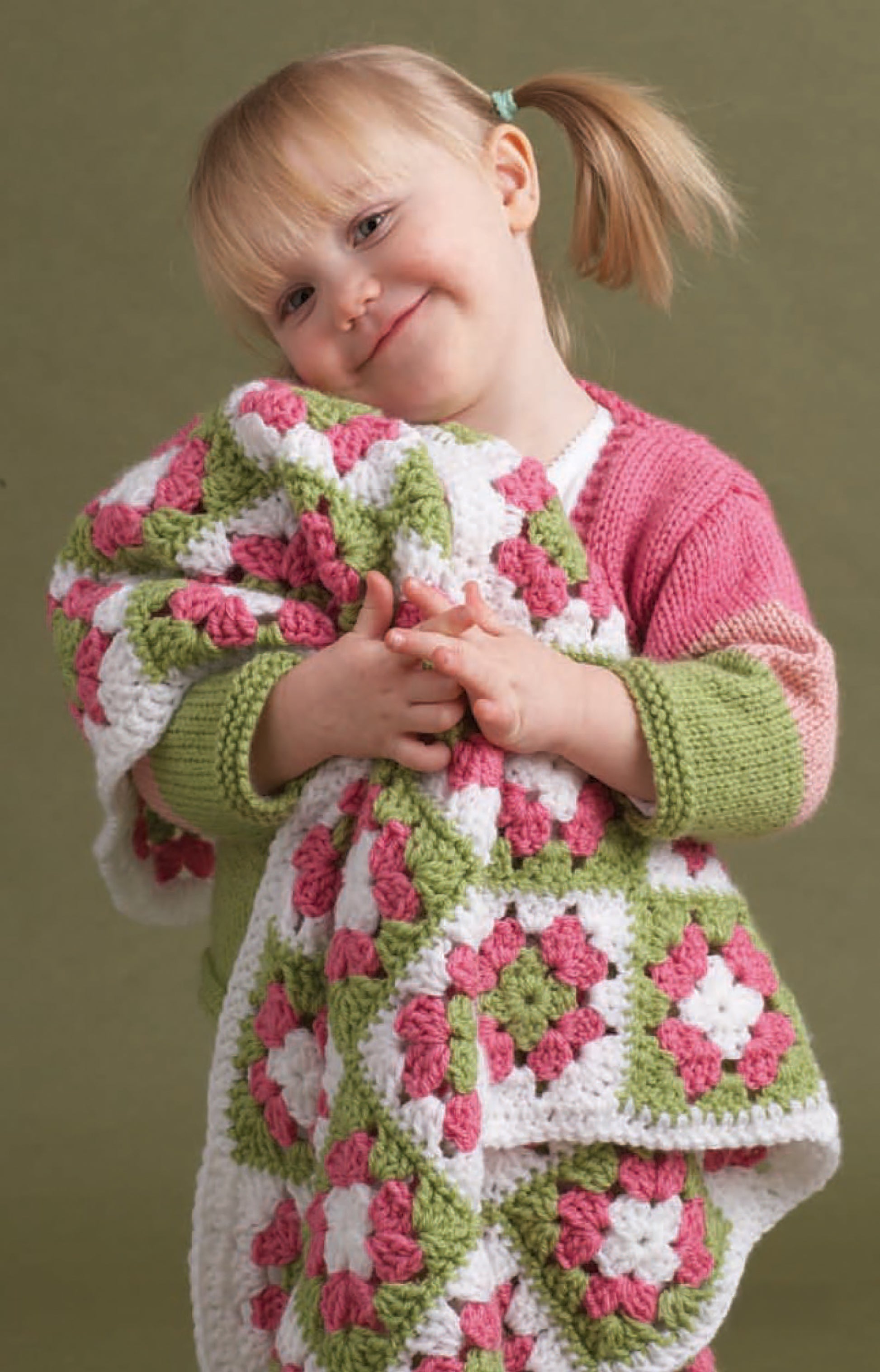 A young child with blonde pigtails smiles while hugging a white, pink, and green granny square blanket crocheted from Vanna's Choice Playful Style by Leisure Arts, against an olive green background.