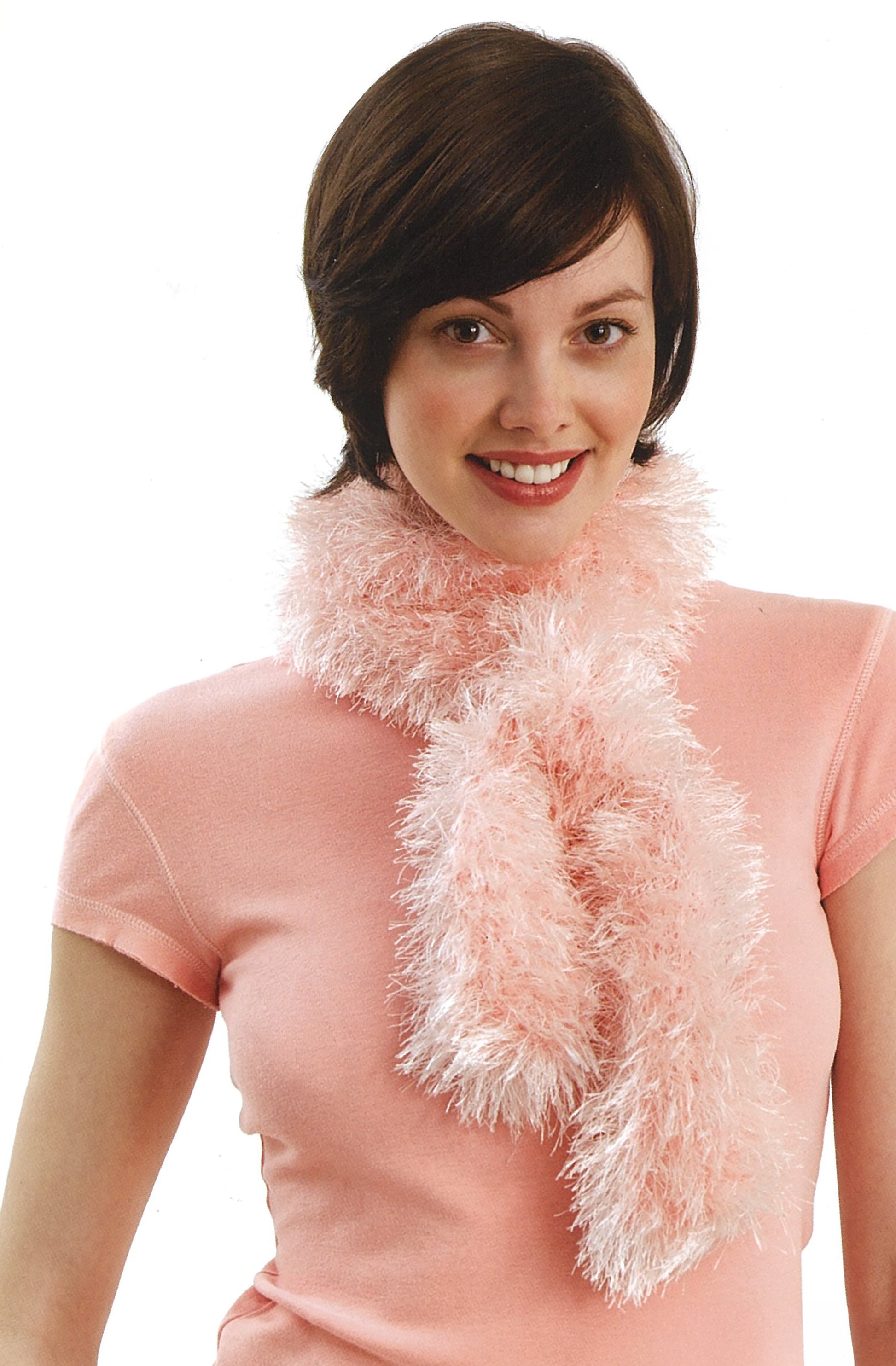A woman with short brown hair smiles while wearing a scarf made from Lion Brand Yarn's Absolutely Fun Fur, featured by Leisure Arts, along with a matching pink short-sleeve shirt against a white background.