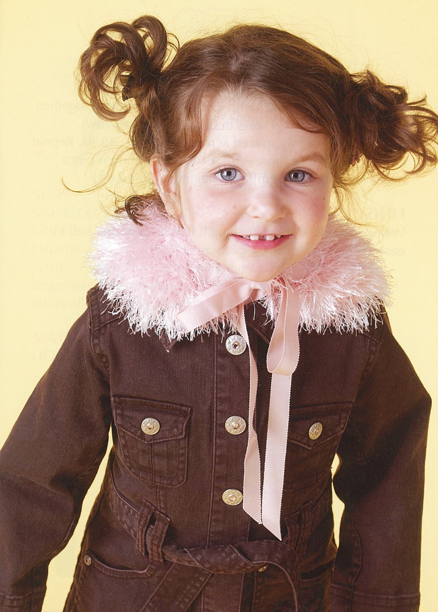 A smiling young girl with brown hair in pigtails wears a brown jacket accented by a fluffy collar made from Lion Brand Yarn's Absolutely Fun Fur, featured by Leisure Arts, standing against a light yellow background.