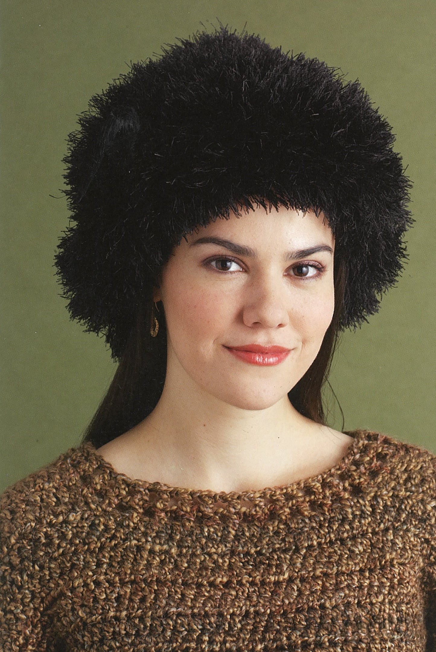 A woman wearing a hat made from Lion Brand Yarn's Absolutely Fun Fur Digital Download by Leisure Arts and a brown textured sweater stands against a plain green background, smiling gently.