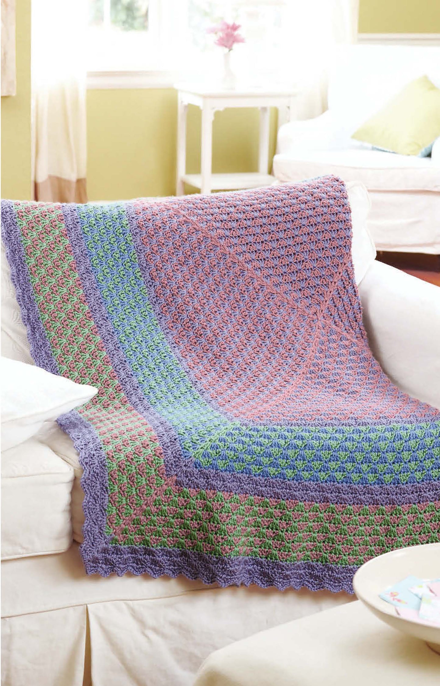 The Grannies Gone Wild crochet afghan by Leisure Arts, featuring vibrant purple, green, pink, and blue sections, adds a lively touch to a white sofa—perfect inspiration for beginner crochet projects in any bright living space.