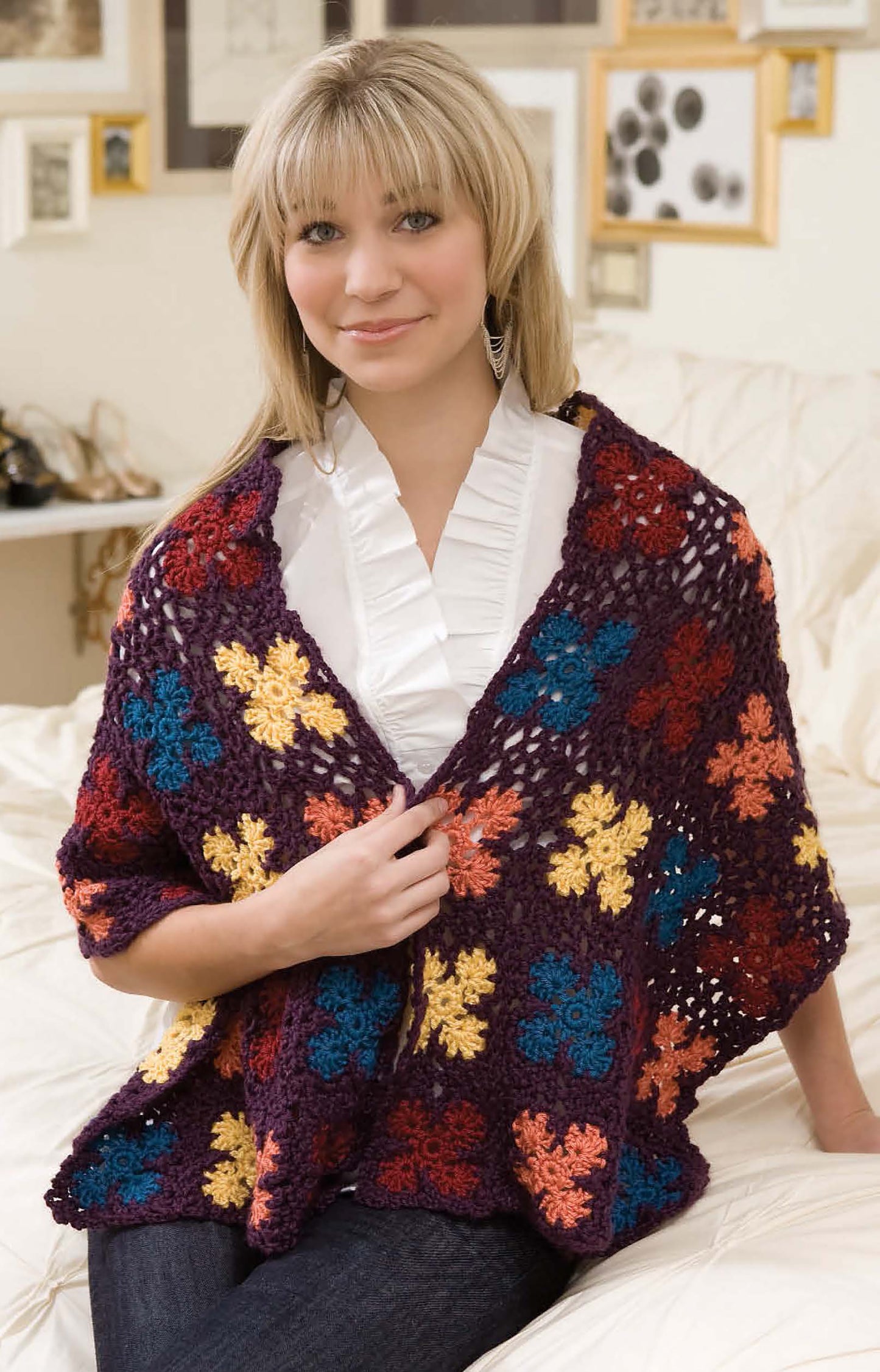 A smiling woman with blonde hair and bangs sits indoors in blue jeans, a white ruffled blouse, and the colorful "Grannies Gone Wild" crochet wrap by Leisure Arts—an ideal beginner project with a floral pattern. Framed art decorates the wall behind her.