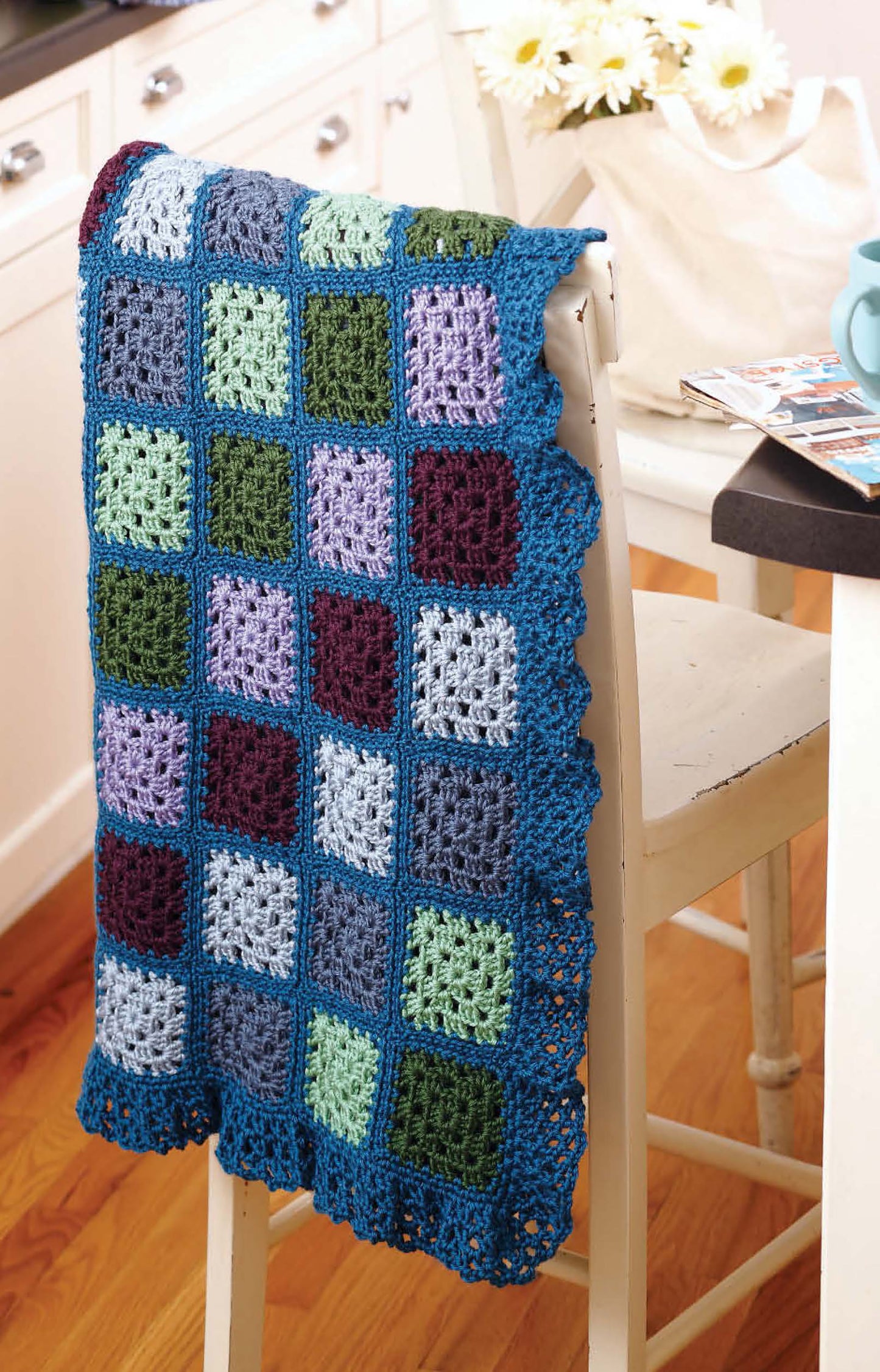 The "Grannies Gone Wild" crochet blanket by Leisure Arts features vibrant granny squares and a scalloped border—perfect for beginners—and is shown draped over a white chair in a cozy kitchen with wood floors.