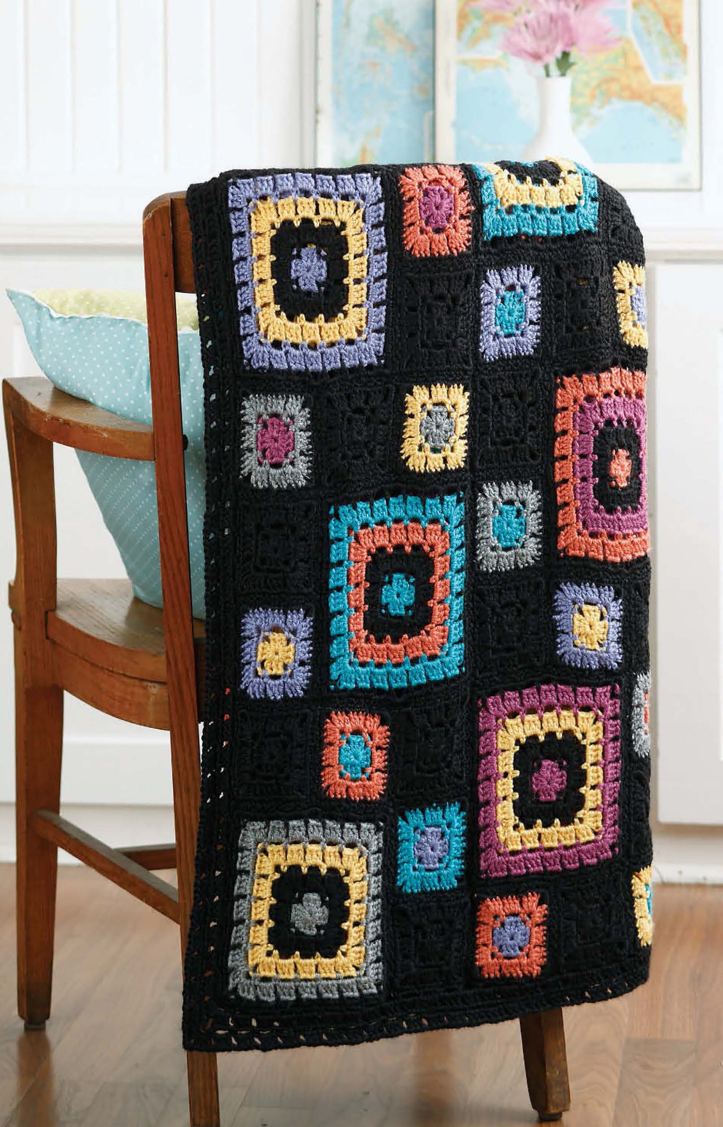The Grannies Gone Wild crochet afghan by Leisure Arts, featuring vibrant multicolored squares edged in black, is draped over a wooden chair with a light blue pillow—ideal inspiration for beginner projects.