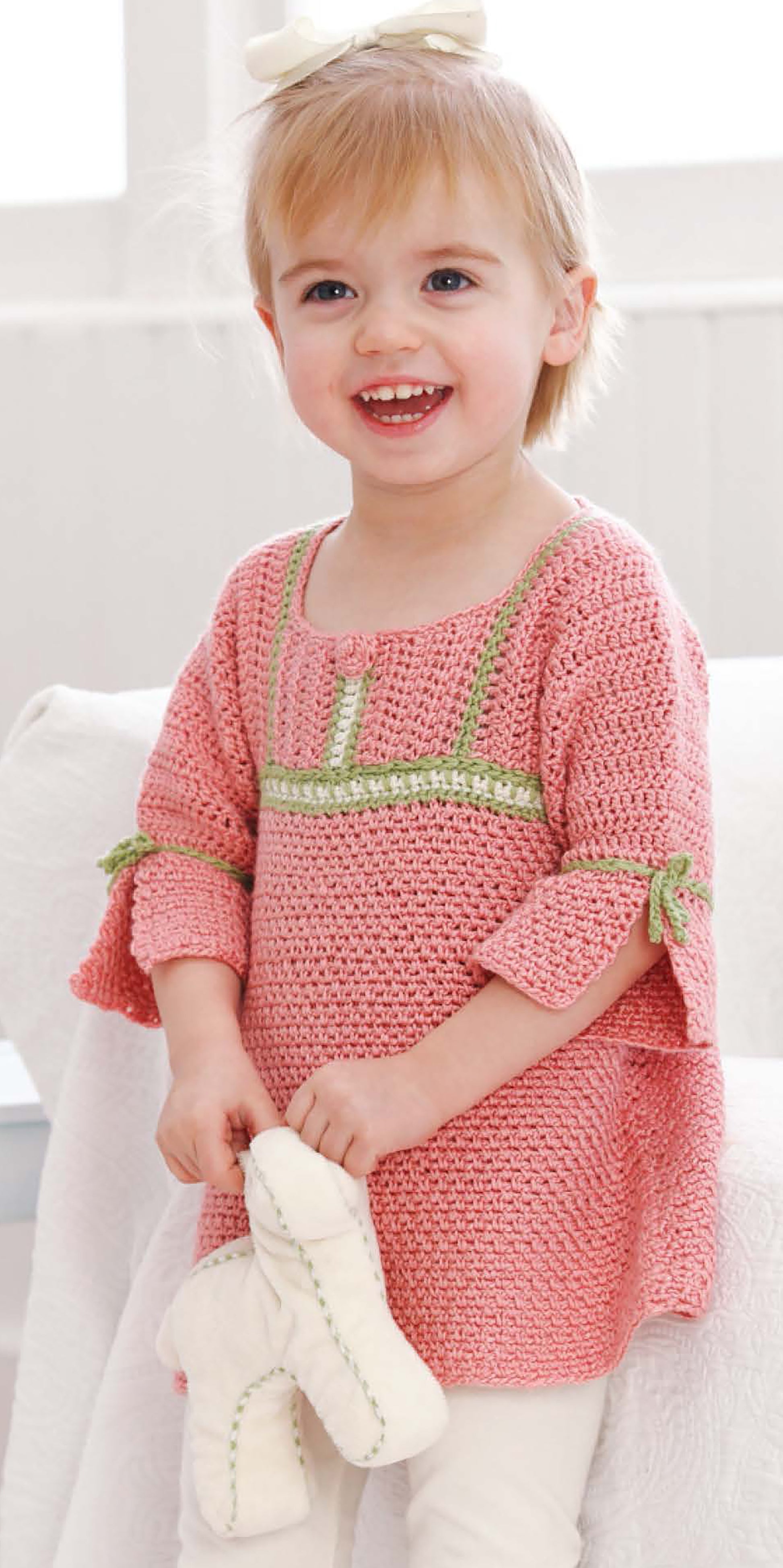 A smiling toddler with short blonde hair models a pink crochet top with green accents from "Sophisticated Babies & Toddlers—6 Crochet Designs Digital Download" by Leisure Arts, while holding white baby shoes.