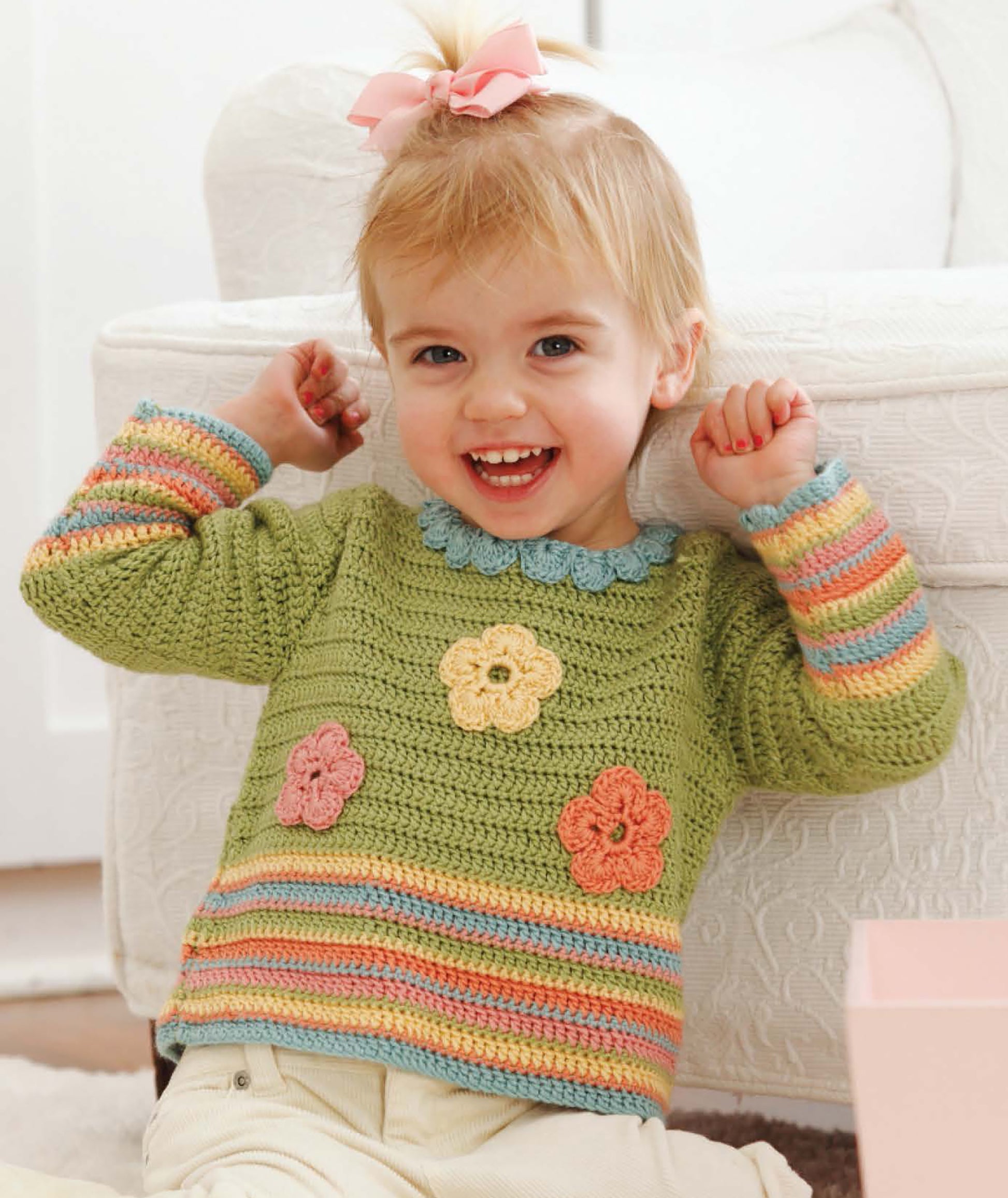 A happy toddler models a green sweater with vibrant crochet patterns from "Sophisticated Babies & Toddlers—6 Crochet Designs Digital Download" by Leisure Arts, sitting with arms raised in a playful pose.