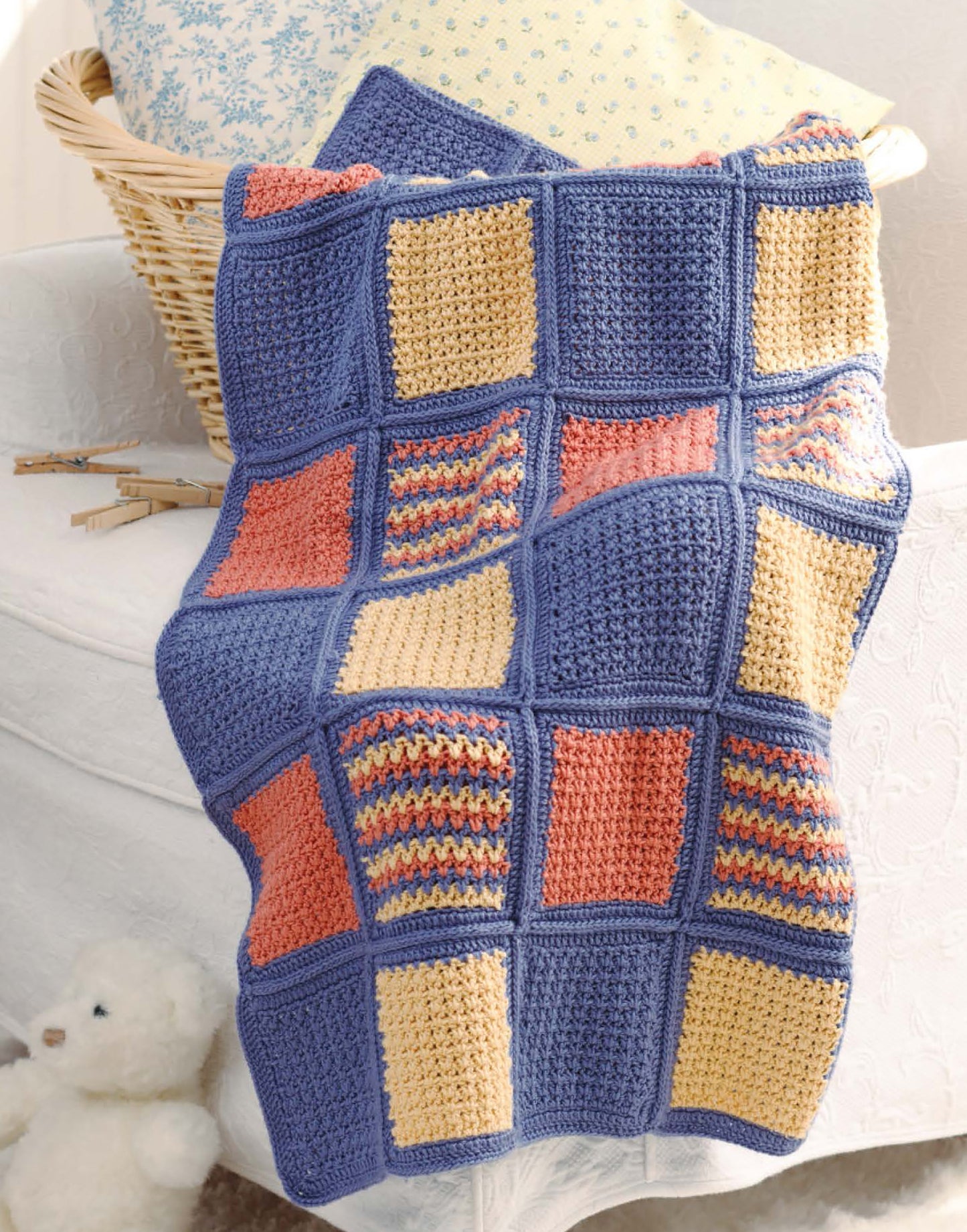 A white chair holds a cozy scene with sweaters, a teddy bear, and a blanket featuring blue borders and colorful squares from the “Sophisticated Babies & Toddlers—6 Crochet Designs” digital download by Leisure Arts.