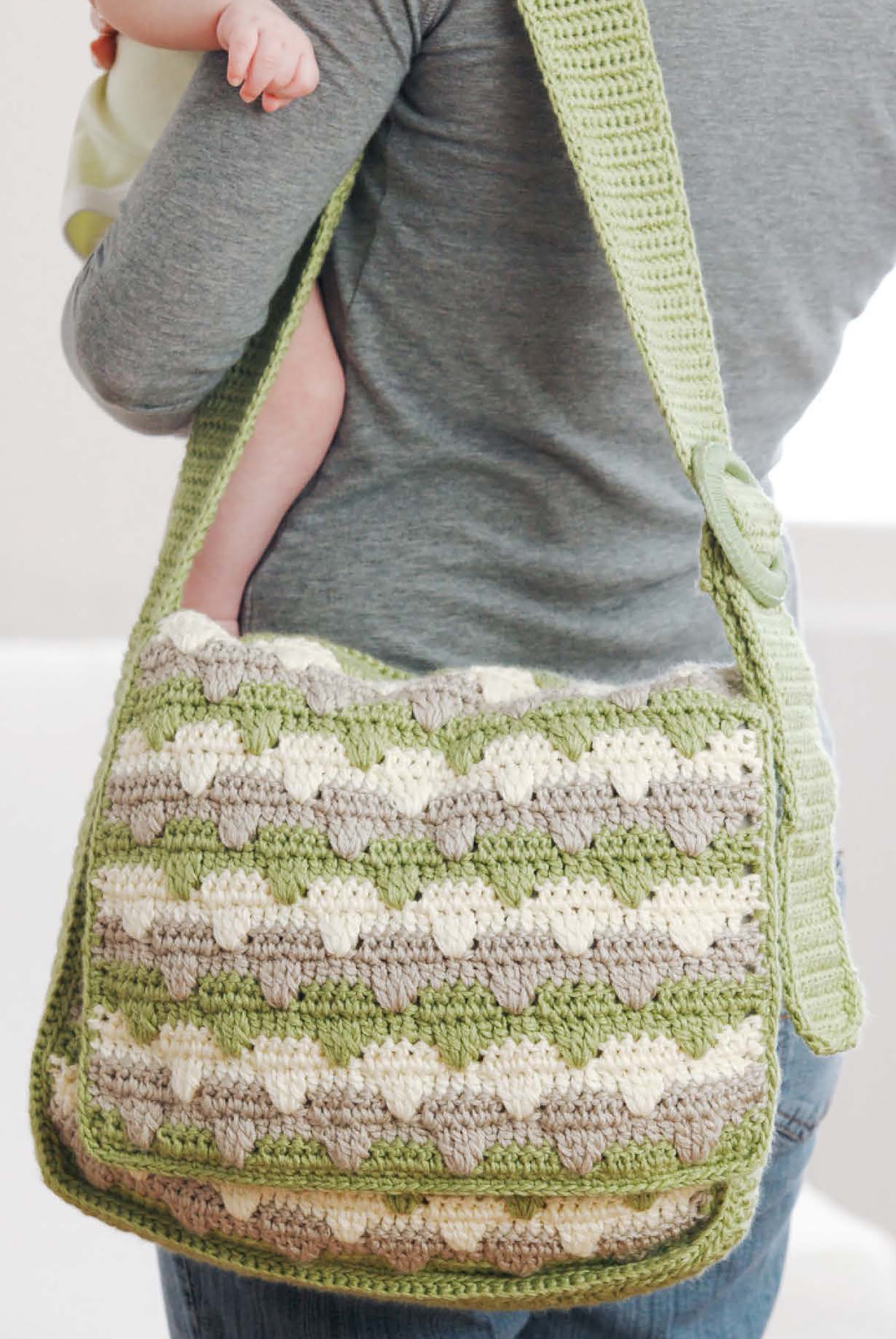 A person in a gray shirt carries a baby and displays a crocheted bag made with green, gray, and cream tones, crafted from "Sophisticated Babies & Toddlers—6 Crochet Designs Digital Download" by Leisure Arts.