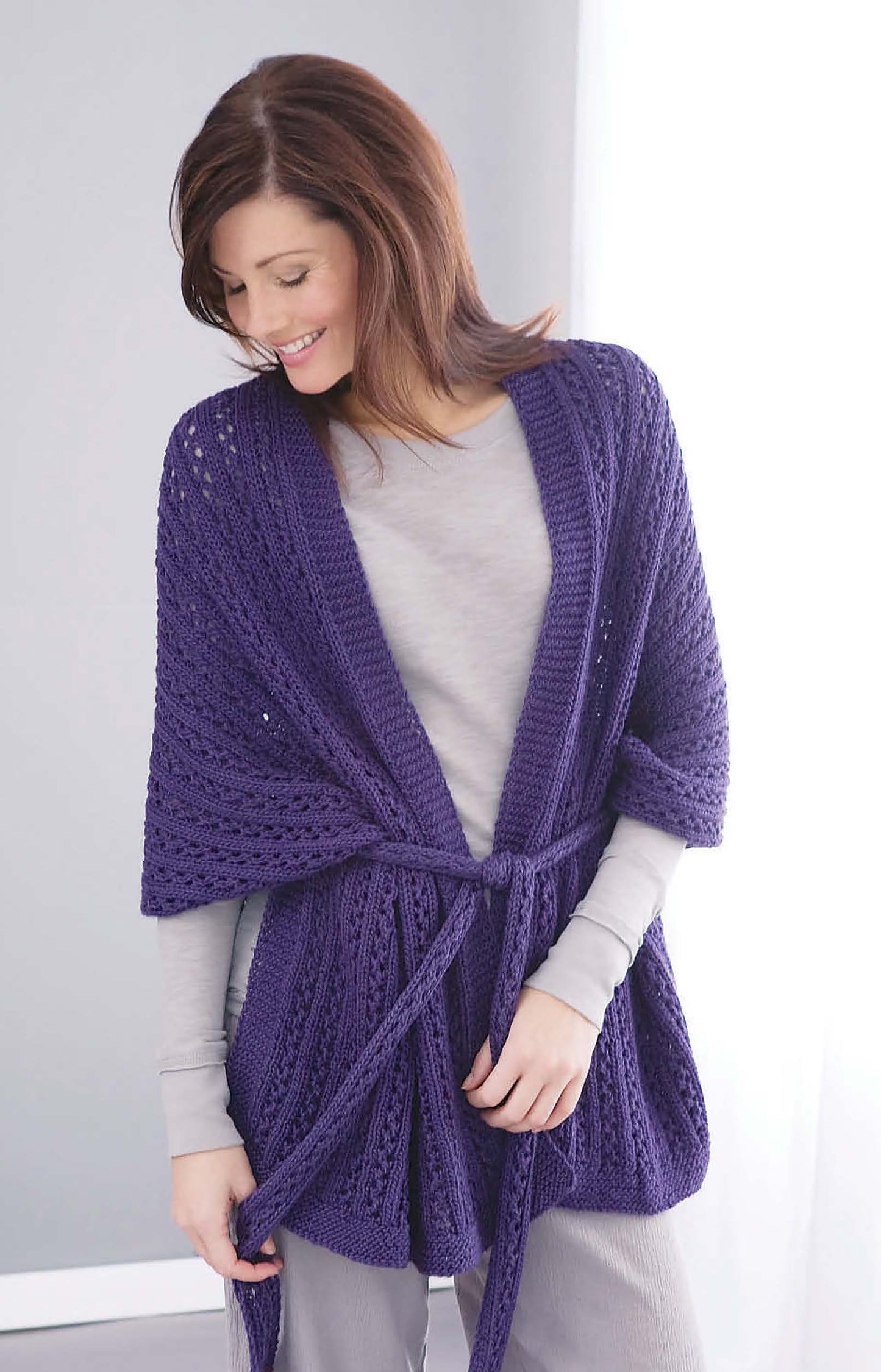 A woman with brown hair wears a long-sleeved gray outfit layered with a deep purple, belted, knitted cardigan made from Caron Country worsted yarn. She models Comfort Knits—6 Knit Designs by Leisure Arts, standing indoors in soft natural light.