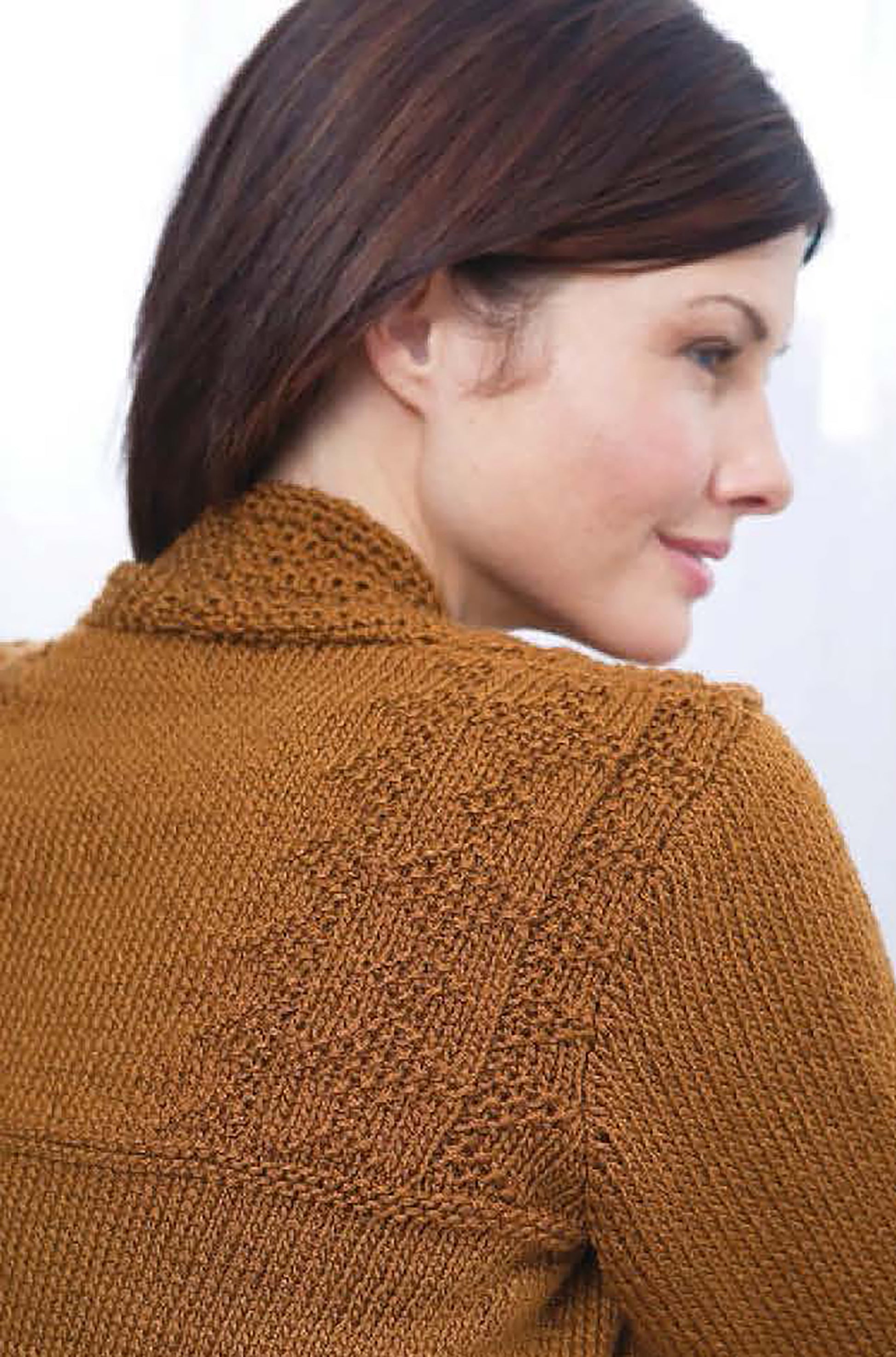 A woman with straight, dark brown hair is seen from the side wearing a textured brown sweater featuring detailed patterns from Comfort Knits—6 Knit Designs; Caron Country by Leisure Arts, crafted in worsted weight yarn on the back and collar.