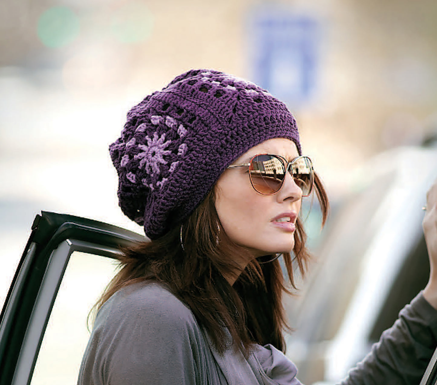 A woman in large sunglasses and a purple Leisure Arts Crochet Celebrity Slouchy Beanie stands by a car door. The blurred background highlights one of our stylish beanies for adults and kids, perfect for any season.