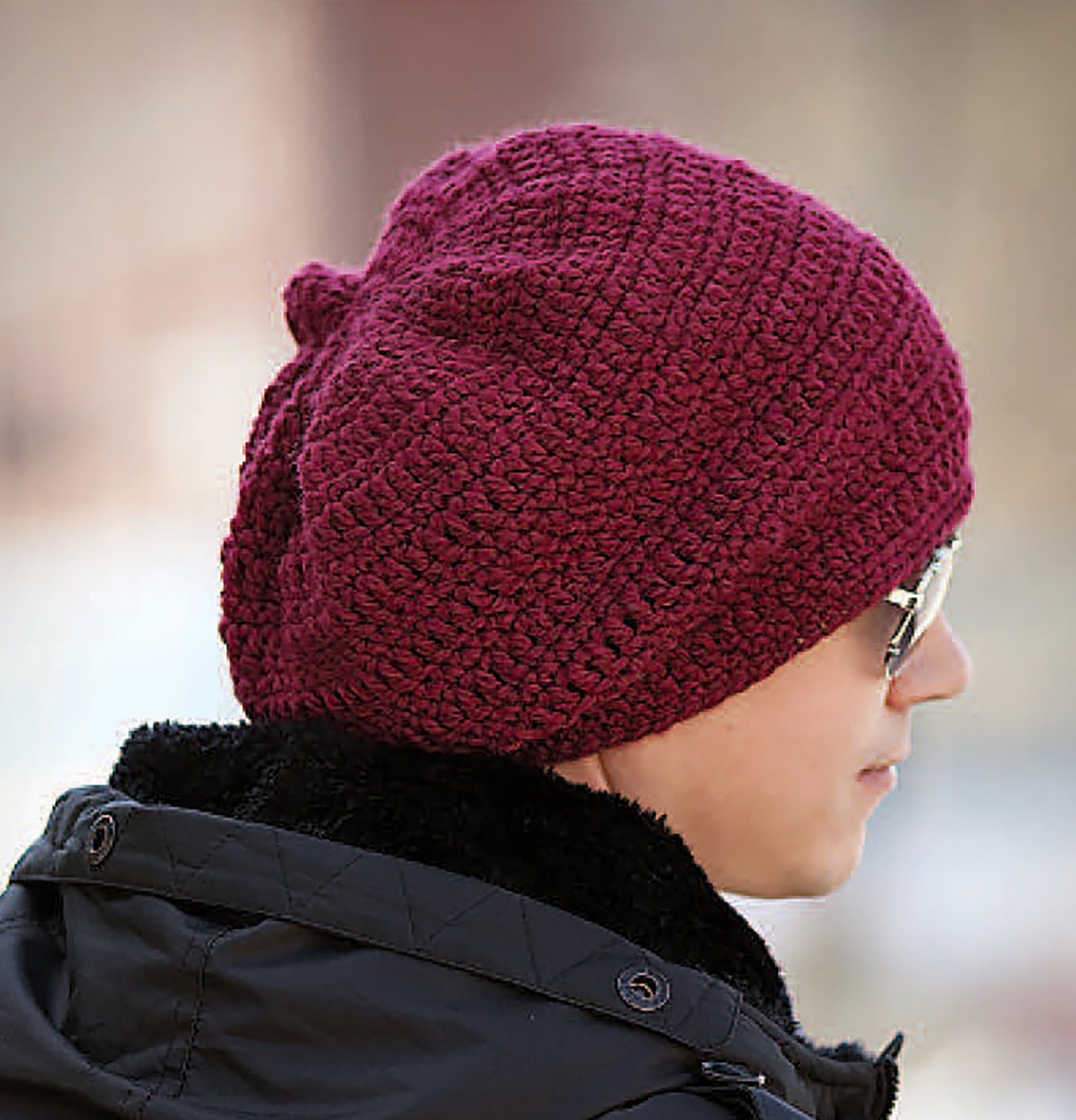 A person in profile wears a maroon knit beanie like the “Crochet Celebrity Slouchy Beanies for the Family” from Leisure Arts, paired with sunglasses and a dark coat, against a blurred outdoor background.