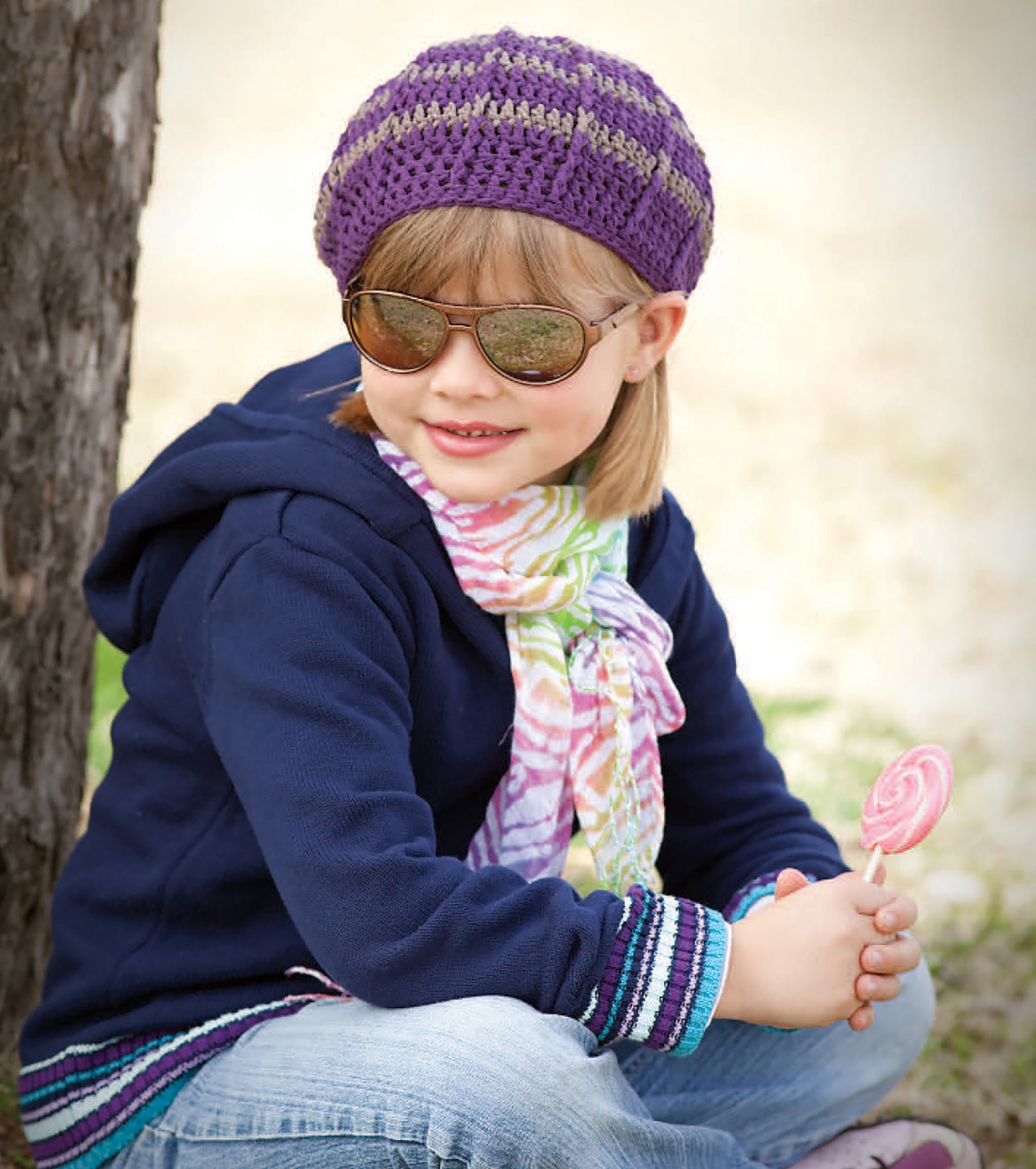 A young child wears Leisure Arts’ Crochet Celebrity Slouchy Beanie, sunglasses, a colorful scarf, and a navy jacket while sitting by a tree outdoors, smiling and holding a pink lollipop.
