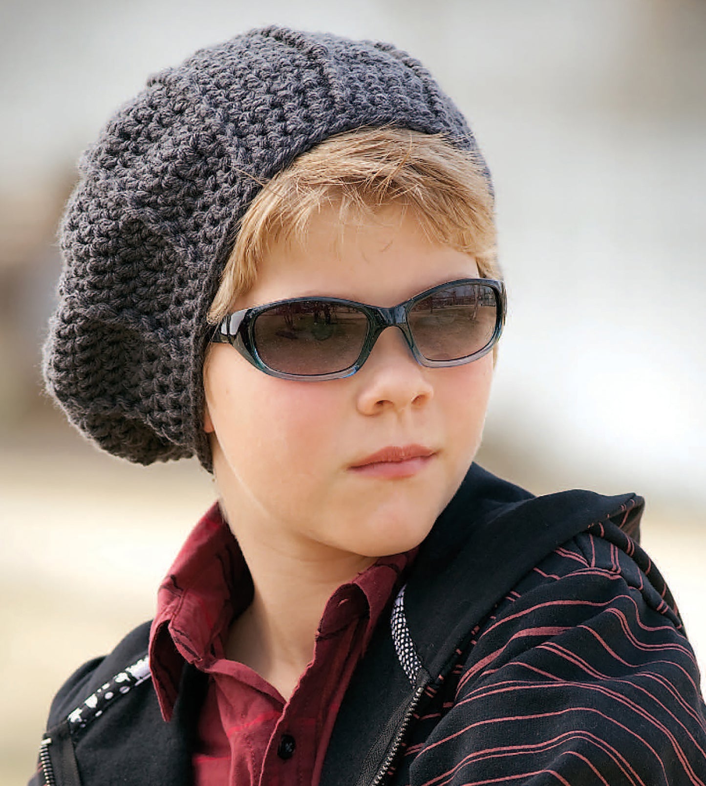 A person wears the Leisure Arts Crochet Celebrity Slouchy Beanie in gray, paired with dark sunglasses and a dark jacket with red stripes, looking to the side with a neutral expression.
