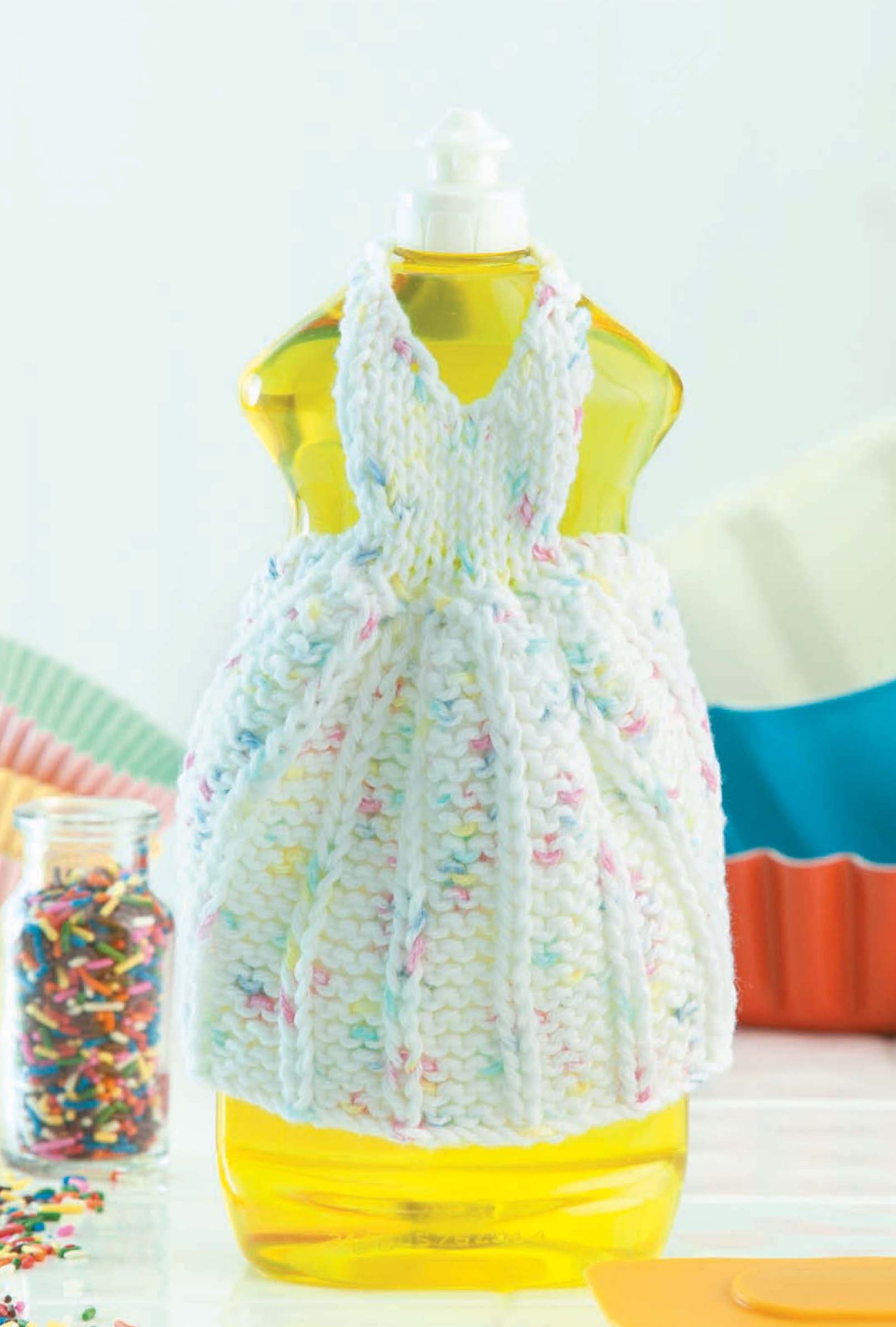 A yellow dish detergent bottle wears a charming, white speckled knitted cover from the Leisure Arts digital pattern "Dishcloth Dresses—The Cutest Quick & Easy Gifts, Ever!" Baking supplies and sprinkles blur in the background.
