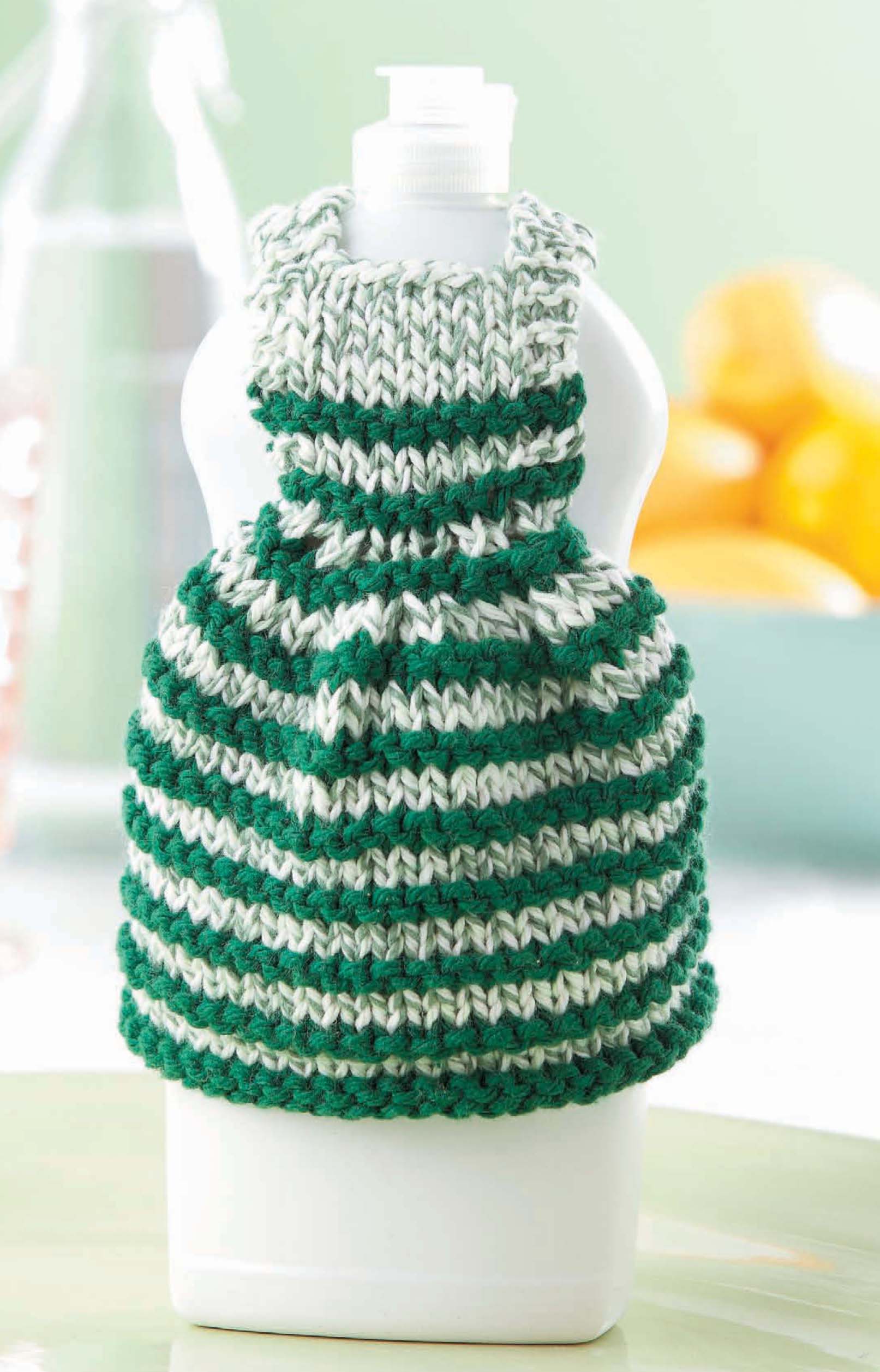 A white detergent bottle is dressed in a green and white striped cozy, made from cotton yarn dishcloth material. Featured: Dishcloth Dresses—The Cutest Quick & Easy Gifts, Ever! Digital Download by Leisure Arts. Blurred lemons appear in the background.