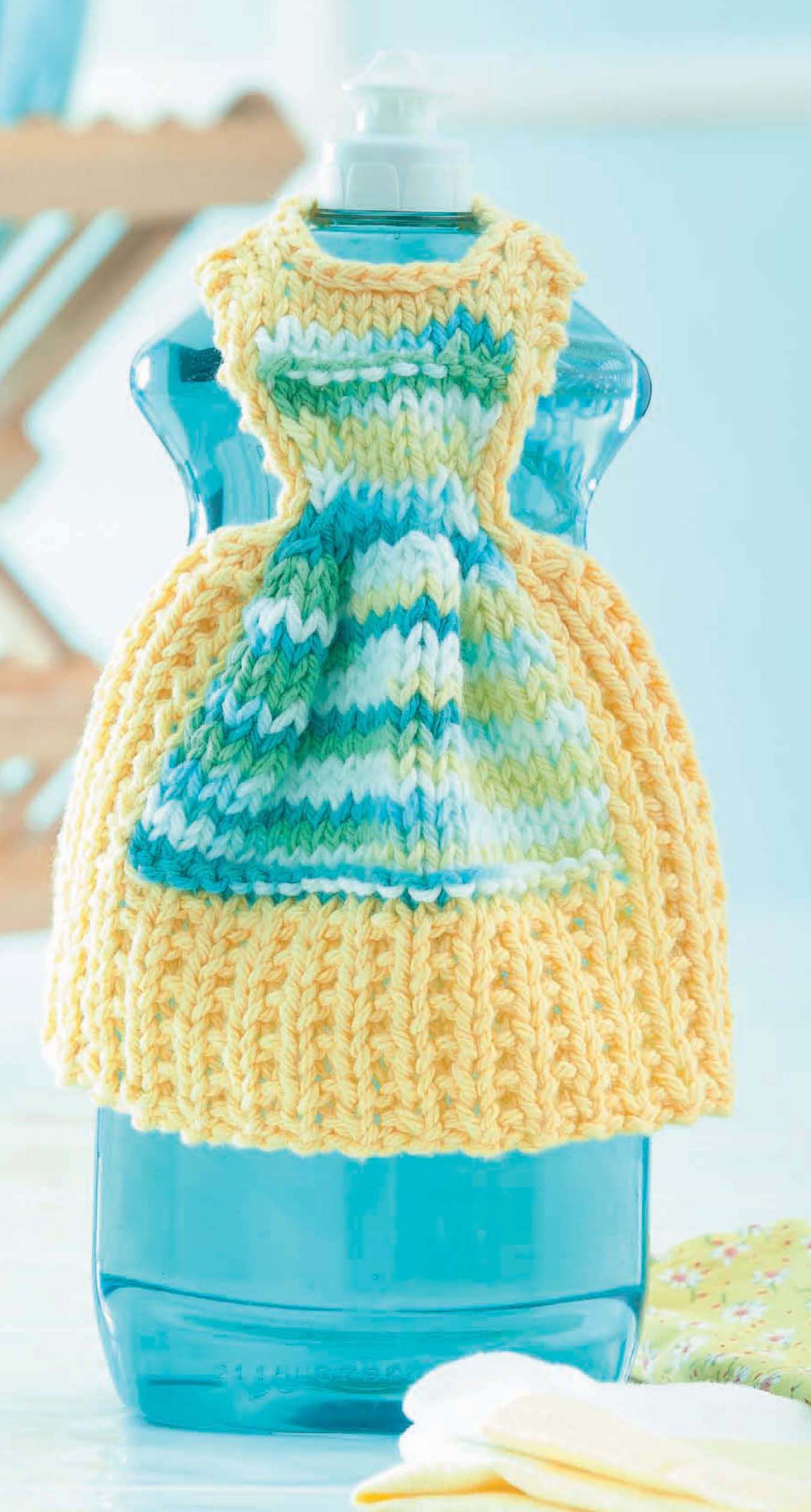 A blue dish soap bottle wears a cozy apron-style dress in yellow, white, blue, and green—made from soft cotton yarn using Leisure Arts' "Dishcloth Dresses—The Cutest Quick & Easy Gifts, Ever!" digital download.